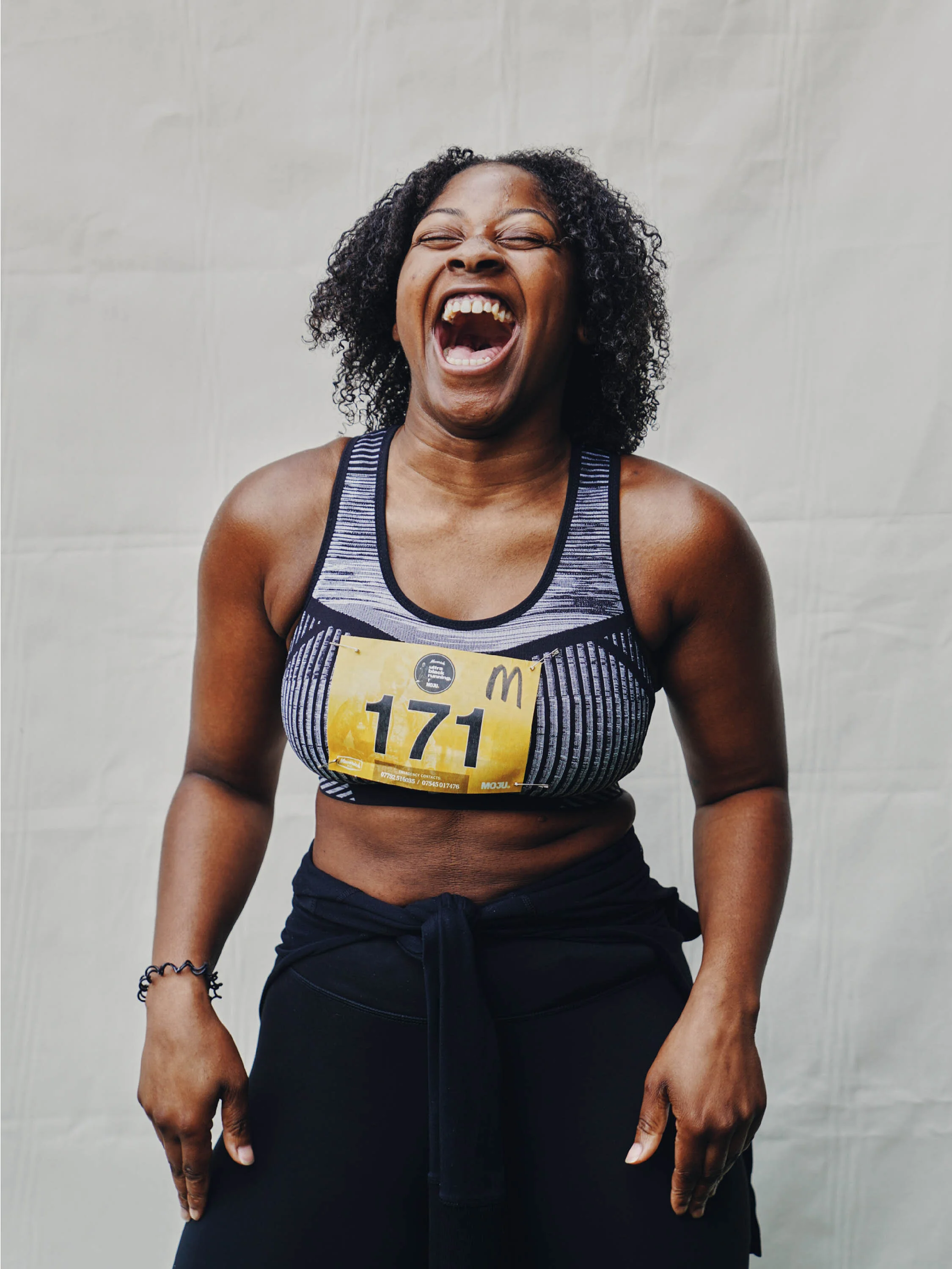 A woman with curly hair laughing, wearing a sports bra with the number 171, and black pants, standing against a plain background.