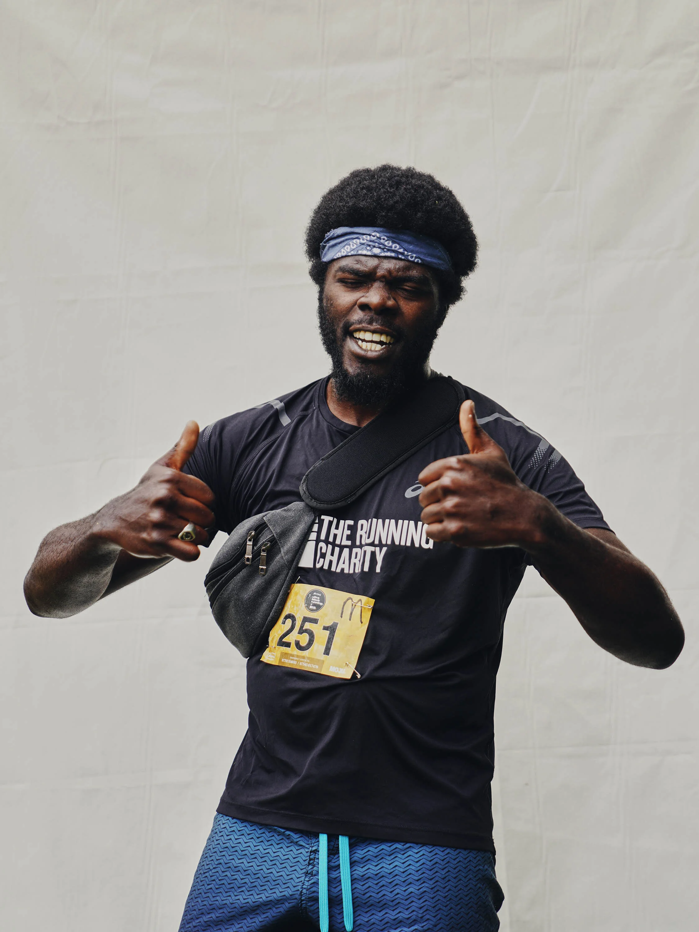 A man wearing a black T-shirt, blue patterned shorts, a blue headband, and a running bib with the number 251, standing against a plain background and giving two thumbs up.