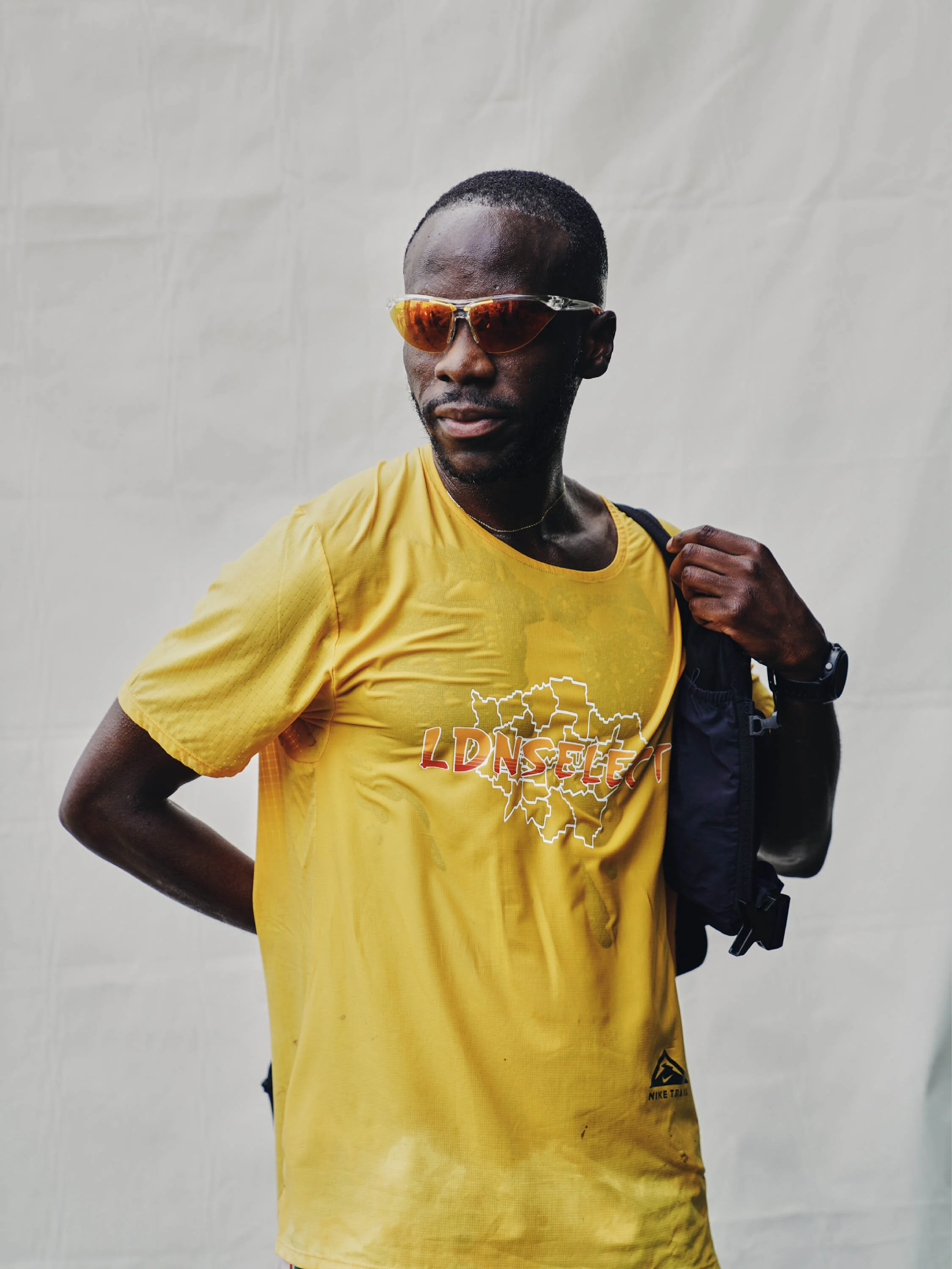 A man with short hair wearing orange sunglasses and a yellow t-shirt with a logo, carrying a backpack.