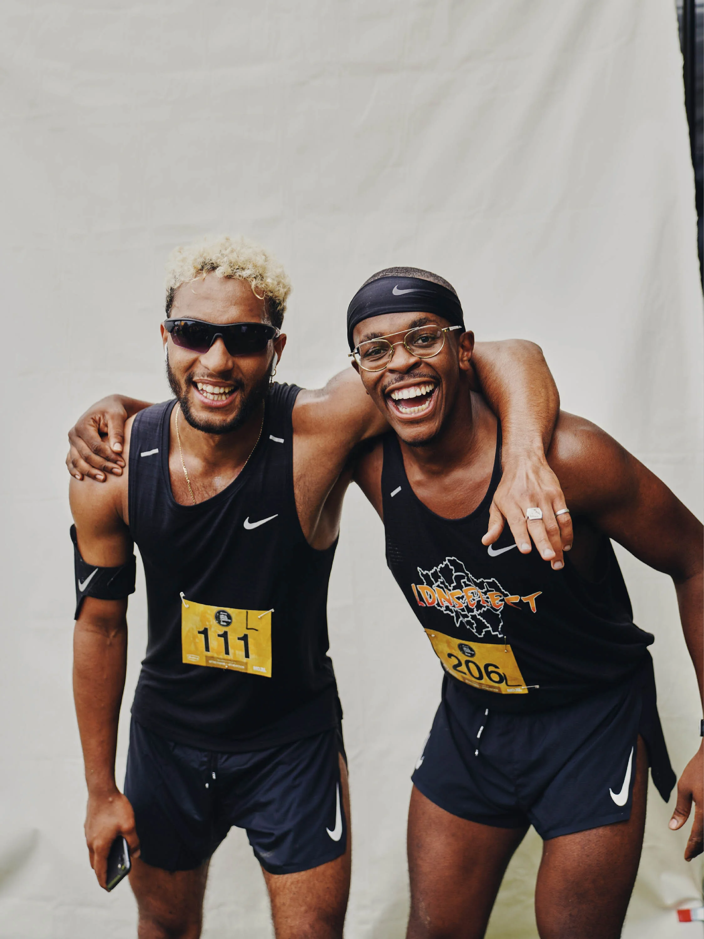 Two male runners in black sportswear smiling and hugging, wearing race bibs, after finishing a race.