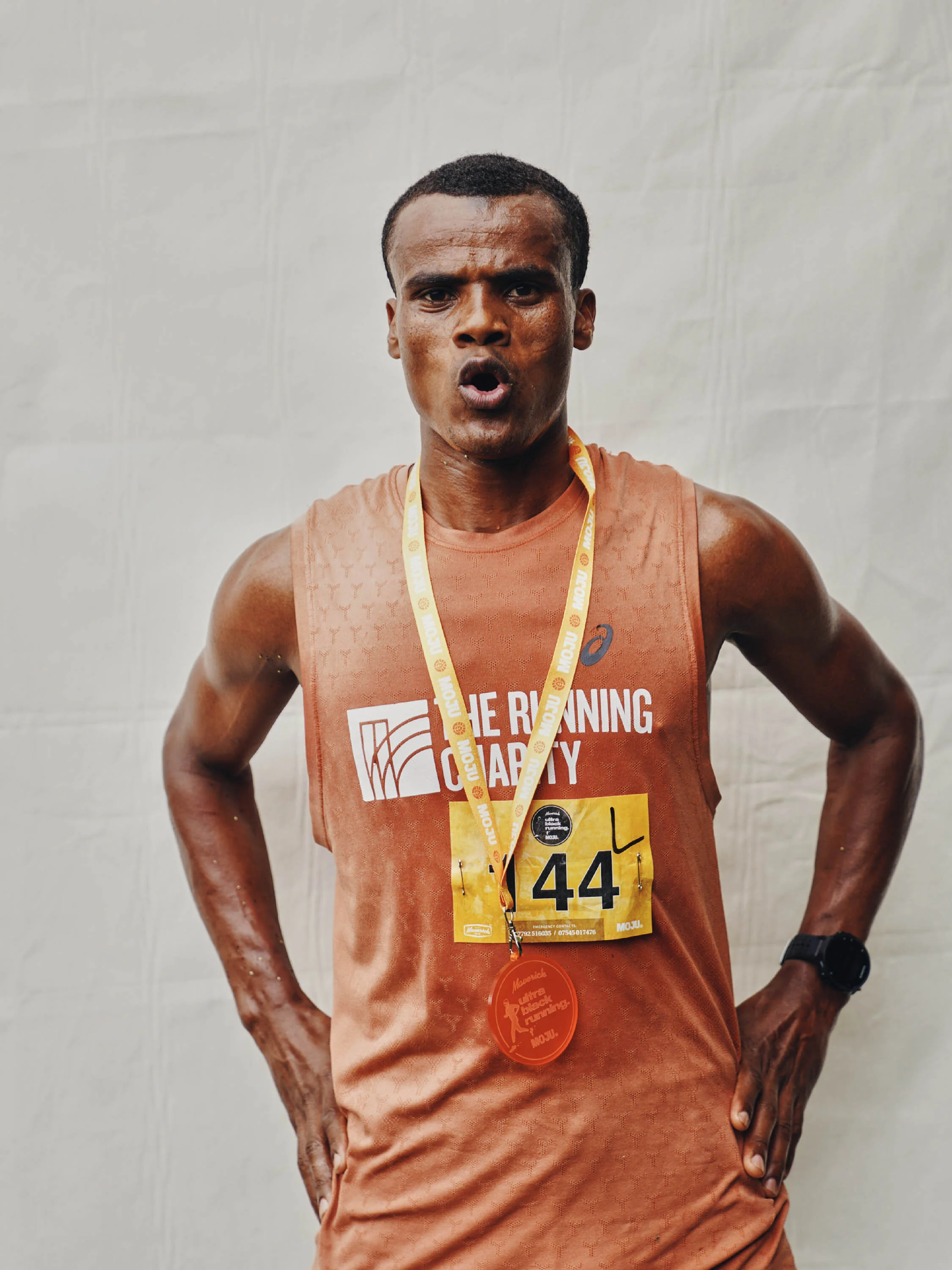 A male marathon runner in orange athletic gear with a medal around his neck, standing with hands on hips, after finishing a race.