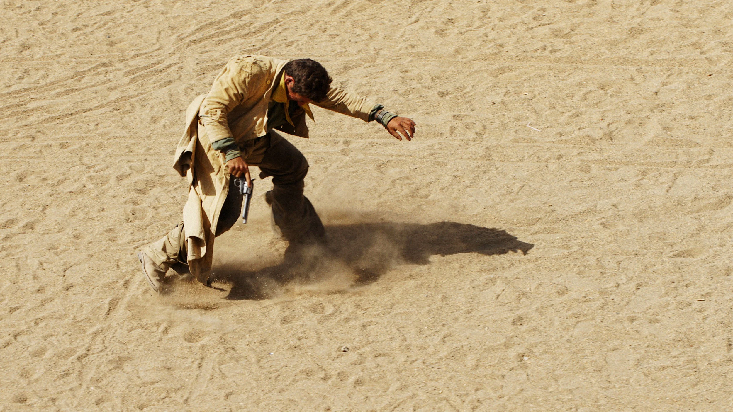 Man in beige jacket and pants crouching in desert, holding a gun, with dust kicking up around his feet.
