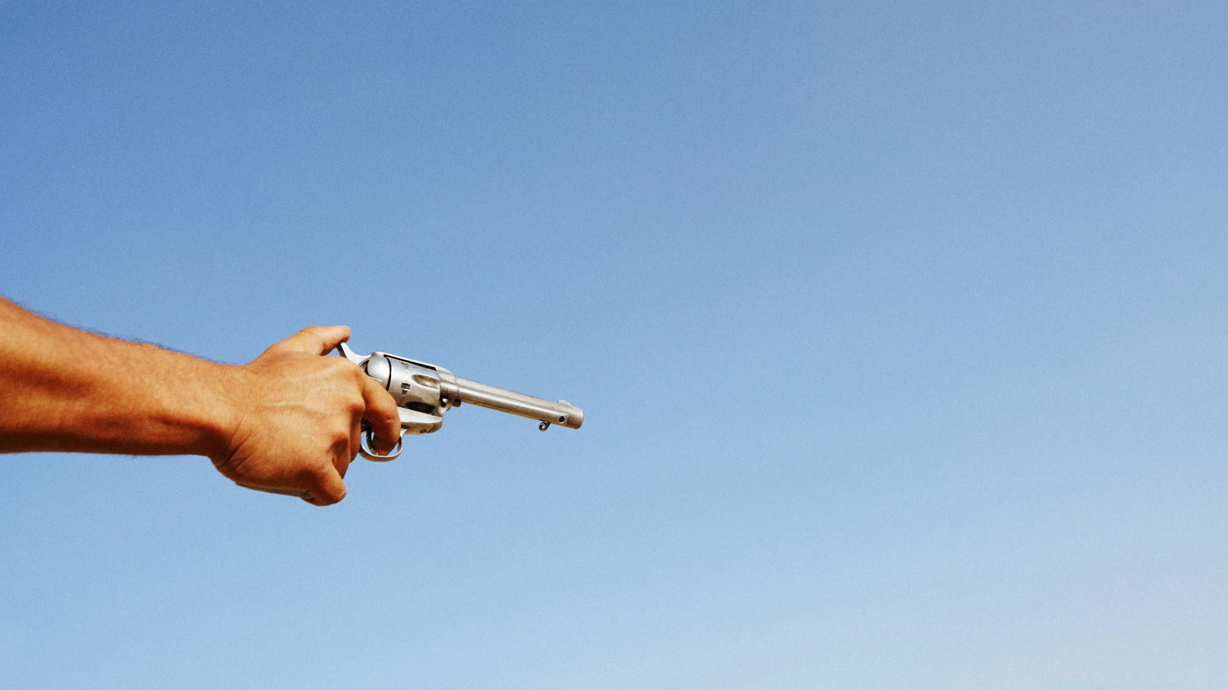 A person's hand holding a silver revolver pointing towards the right against a clear blue sky.