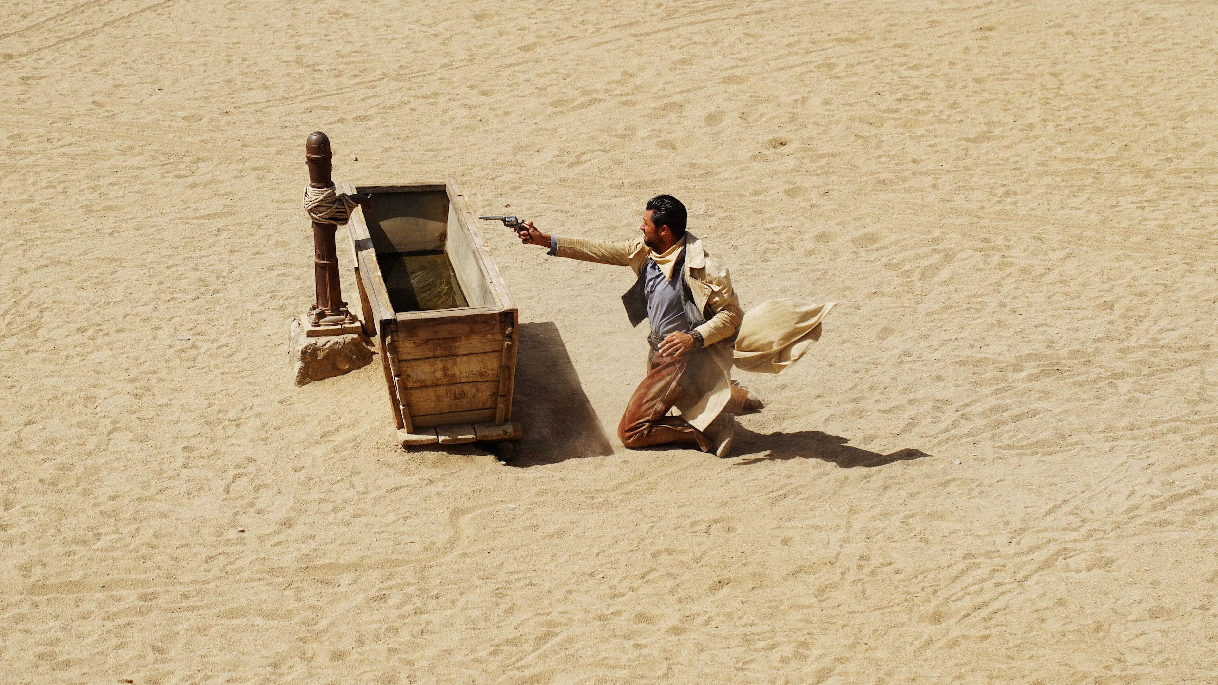 Kneeling man in desert shoots handgun towards water tank, sand surrounds him.
