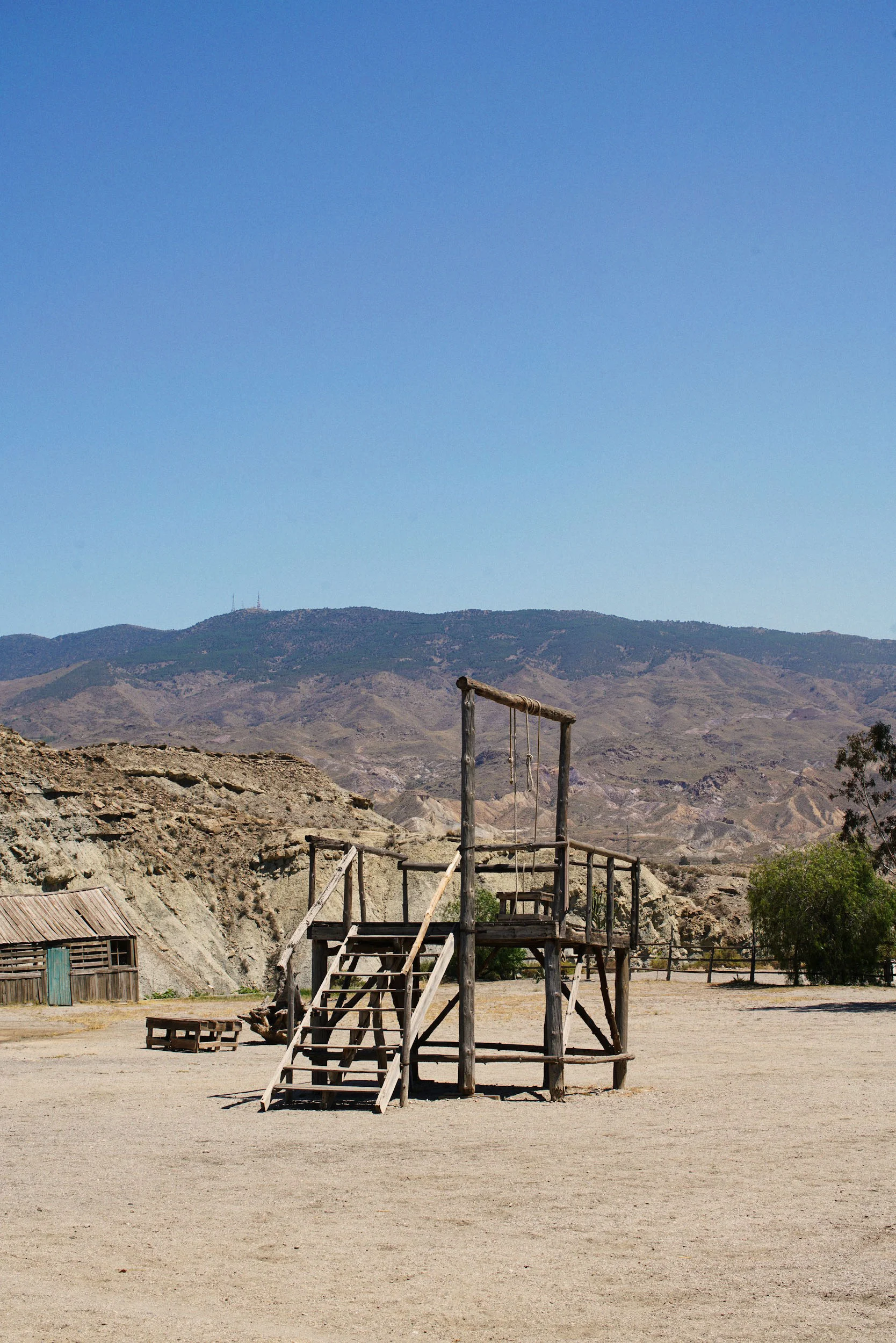 An old wooden playground structure with stairs and a swing set, situated in a desert landscape with mountains in the background.