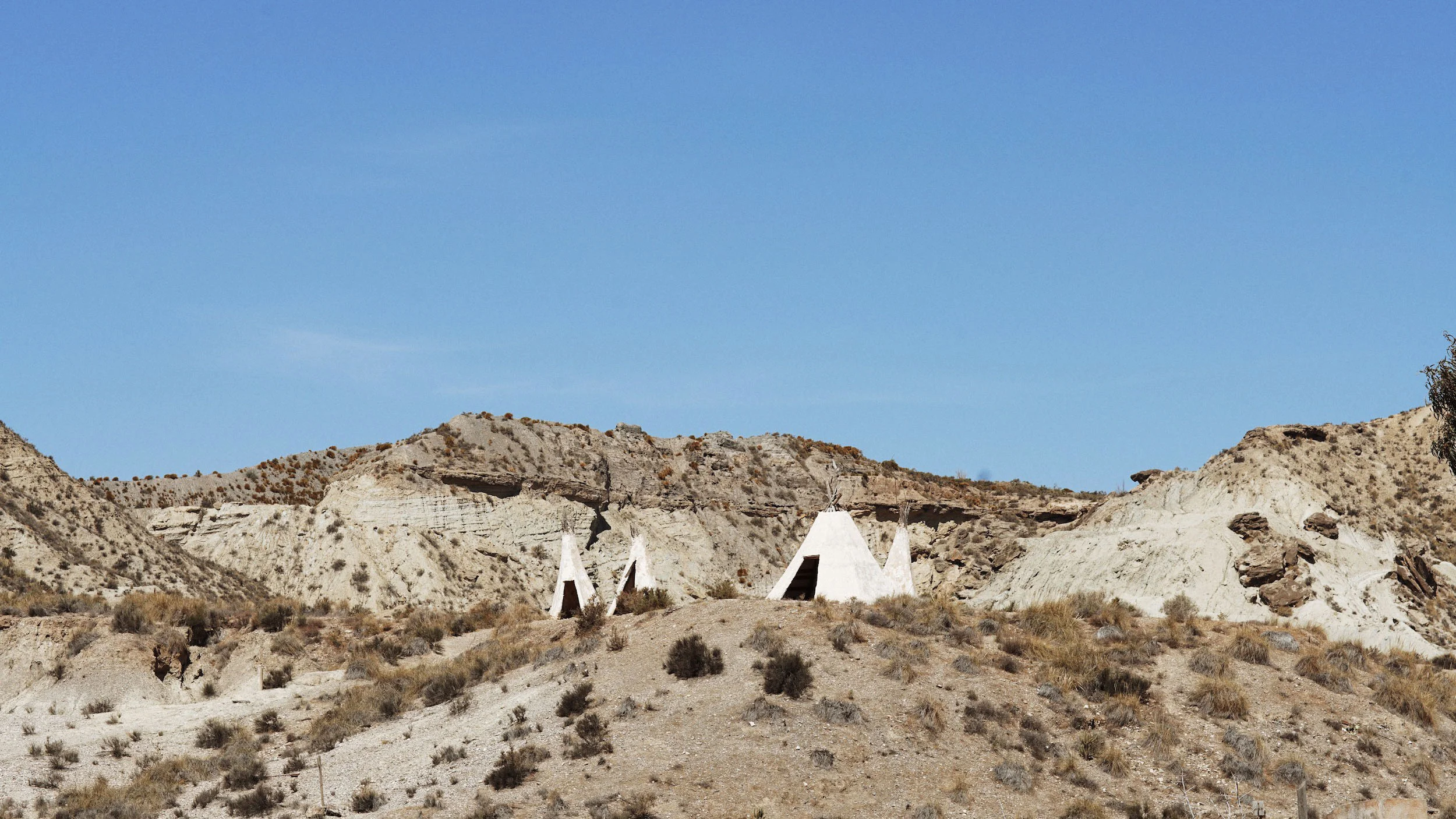 A desert landscape with white Adobe-style triangular structures and rocky hills in the background under a clear blue sky.