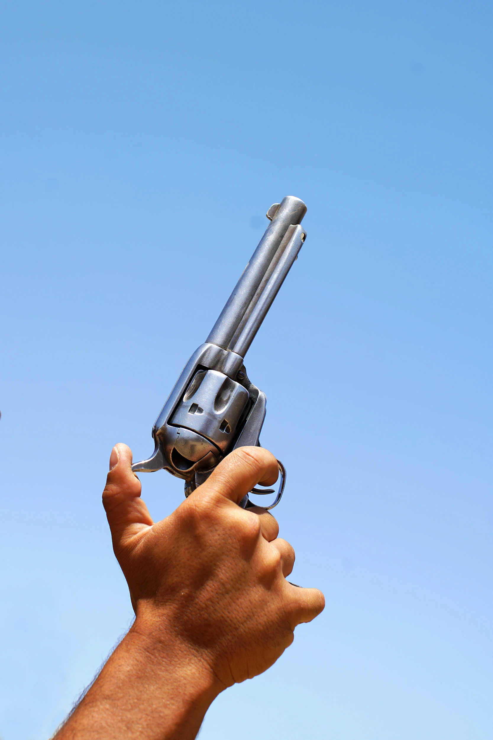 A person's hand holding a silver revolver handgun with the barrel pointed upward against a bright blue sky.