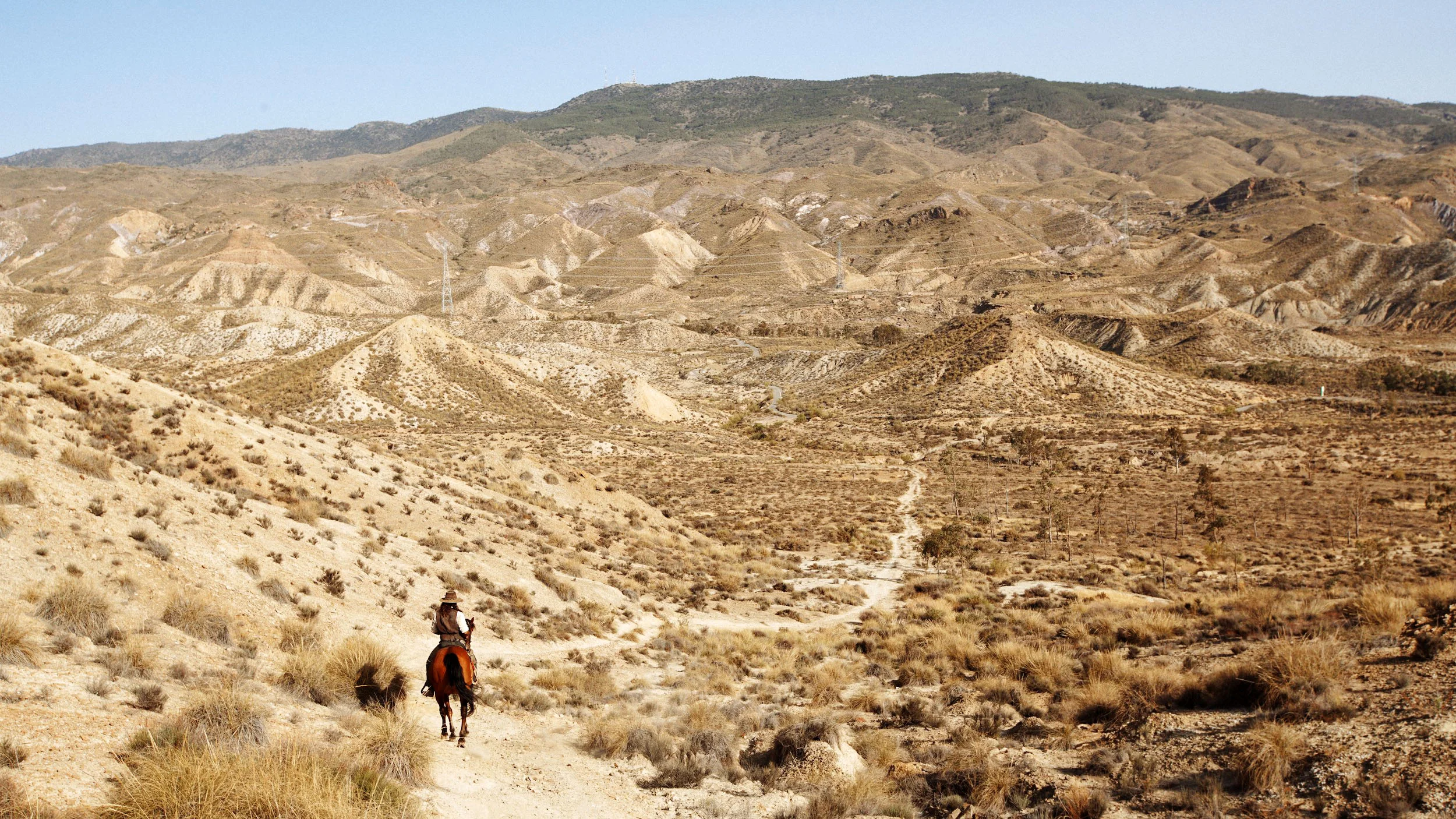 A person riding a horse on a dirt trail through a dry, hilly desert landscape with sparse vegetation and distant mountains.