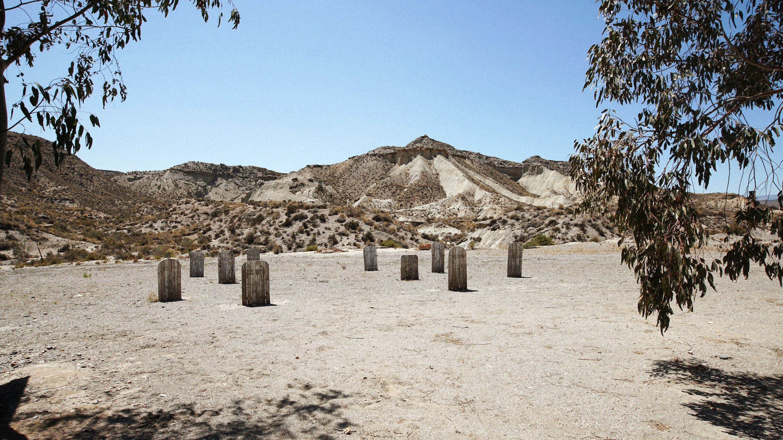 Desert landscape with sandy ground, some wooden posts arranged in a semi-circle, sparse dry vegetation, distant rocky hills, and a clear blue sky.