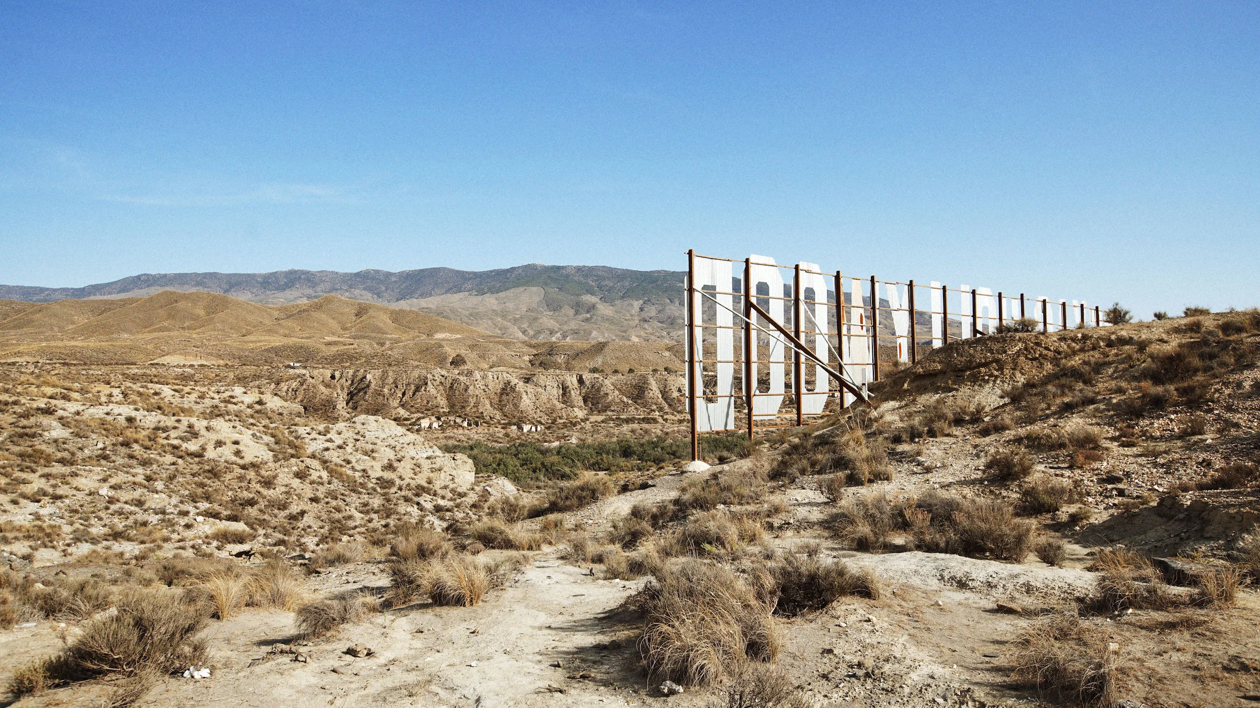 Partially constructed Hollywood sign in a dry, desert landscape with distant hills and mountains under a clear blue sky.