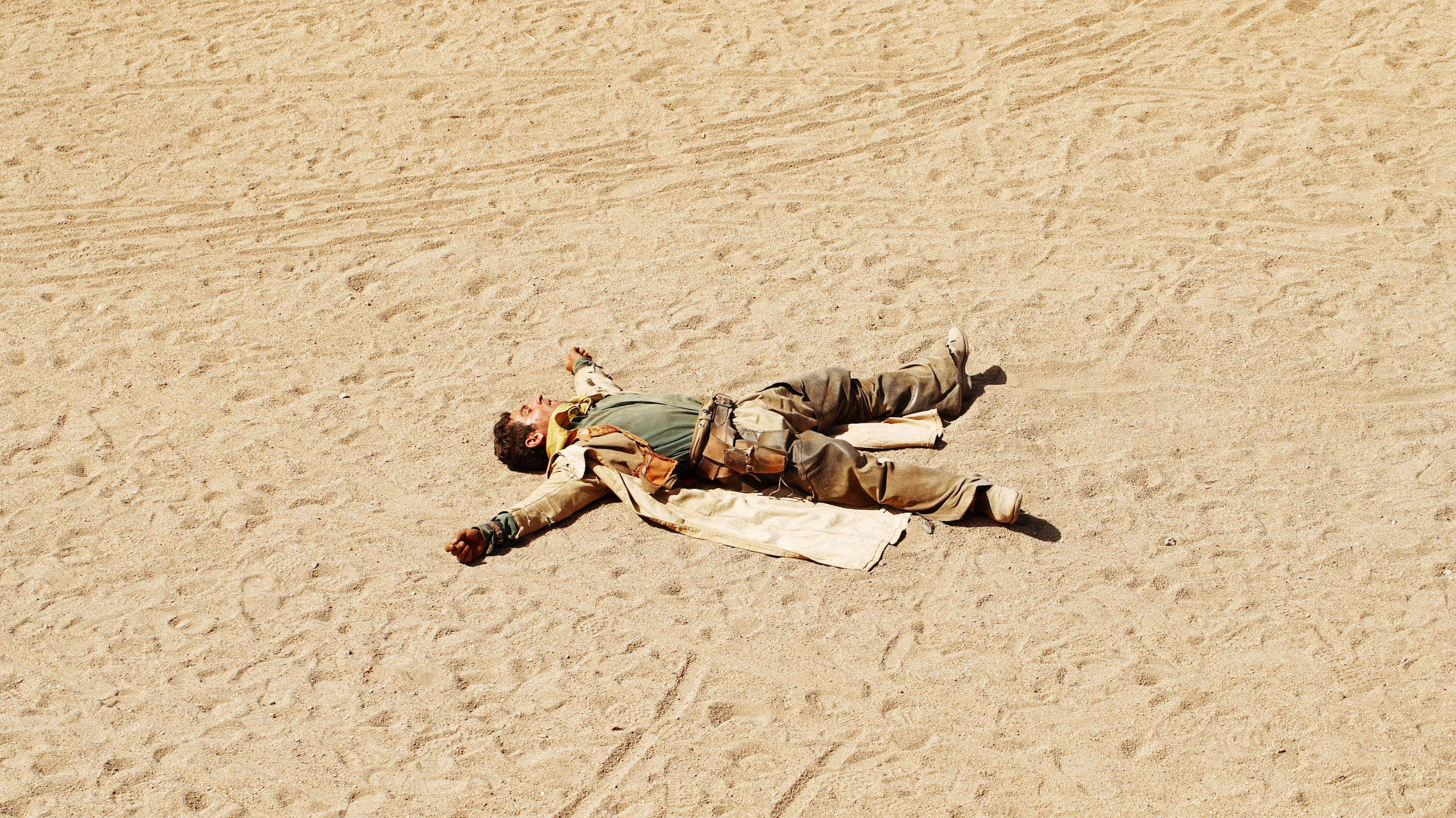 A person lying on the sandy desert ground with arms and legs spread out, appearing to be resting or potentially unconscious.