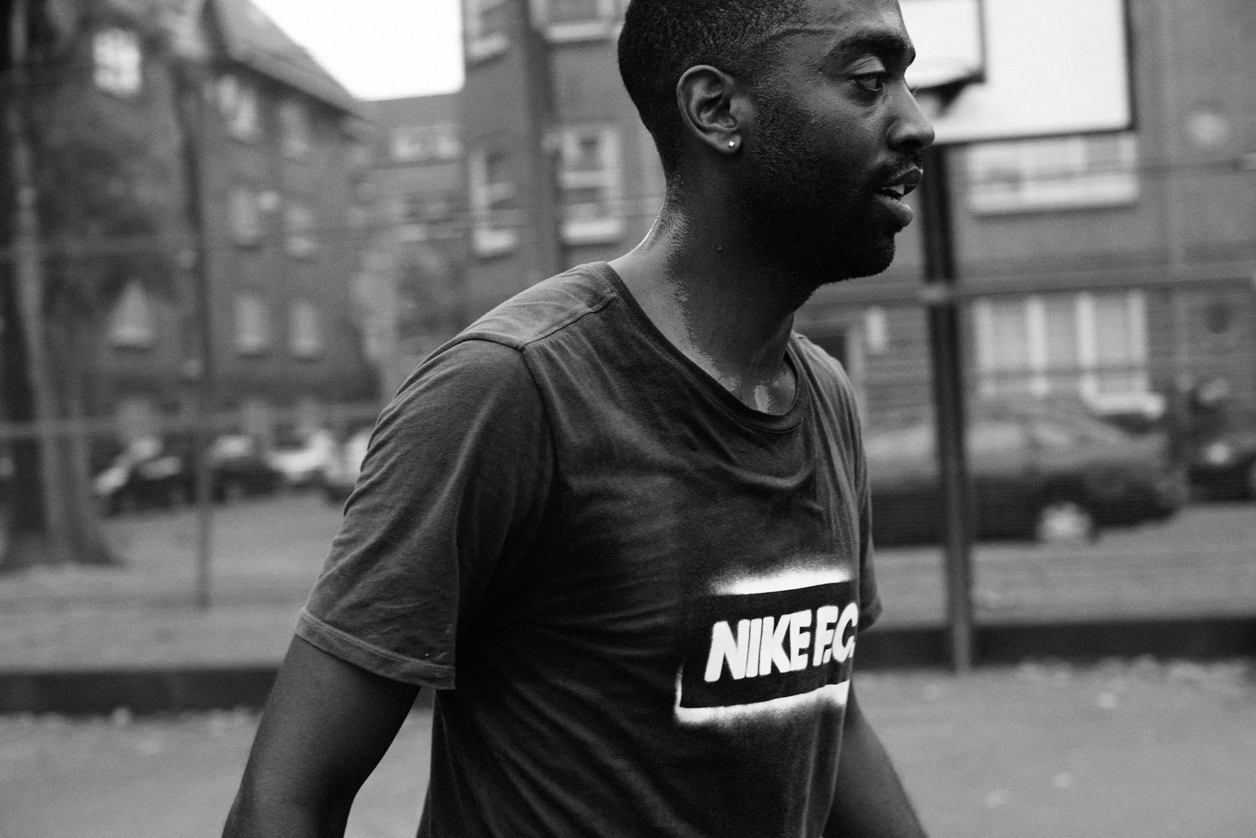 A young man jogging outdoors on a city street, wearing a Nike t-shirt, with buildings and parked cars in the background.
