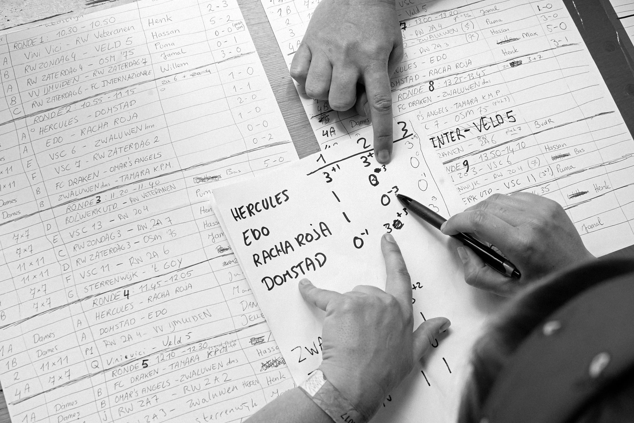 Two people reviewing handwritten sports match scores and schedules on printed and handwritten sheets of paper in black and white.