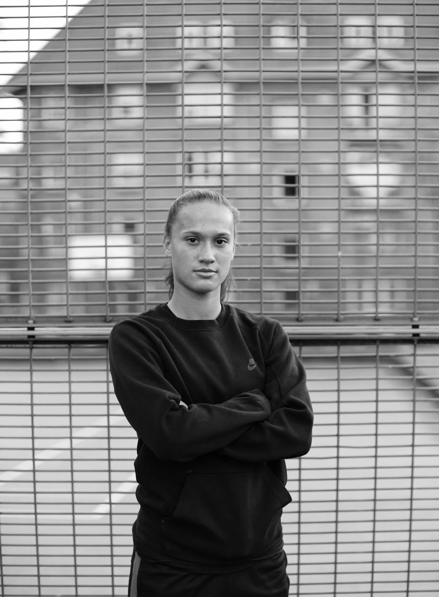 A young woman with light skin and blonde hair tied back, wearing a Nike sweatshirt, standing with arms crossed in front of a wire fence with a residential building in the background.