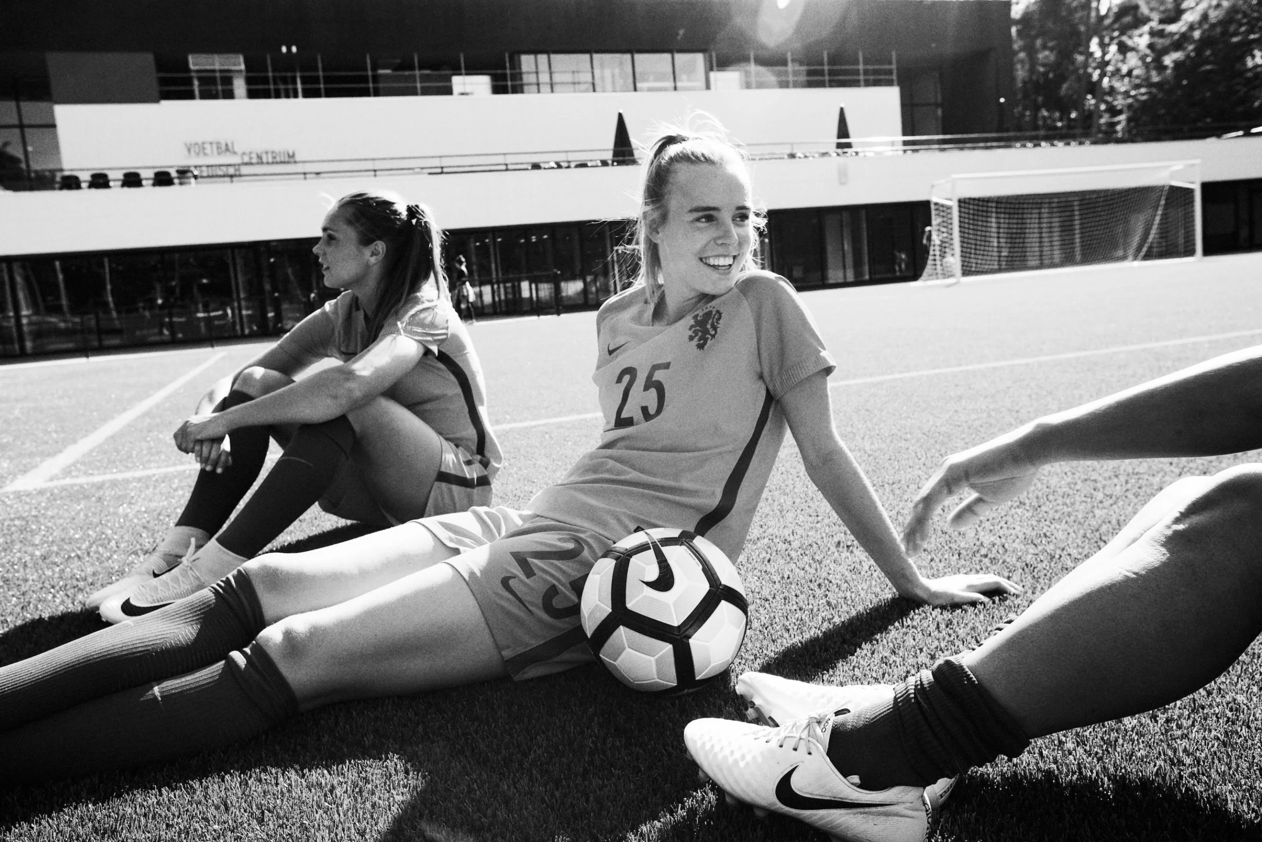 Three female soccer players sitting on the grass on a soccer field, smiling and relaxing after a game, with one girl holding a soccer ball, in front of a modern building with the sign 'VOETBAL CENTRUM'.