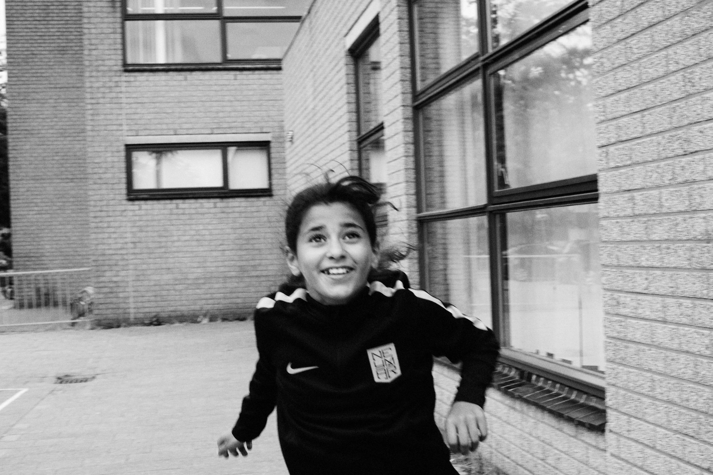 A young boy running outdoors beside a brick building, smiling and looking ahead, with his hair flowing as he moves.
