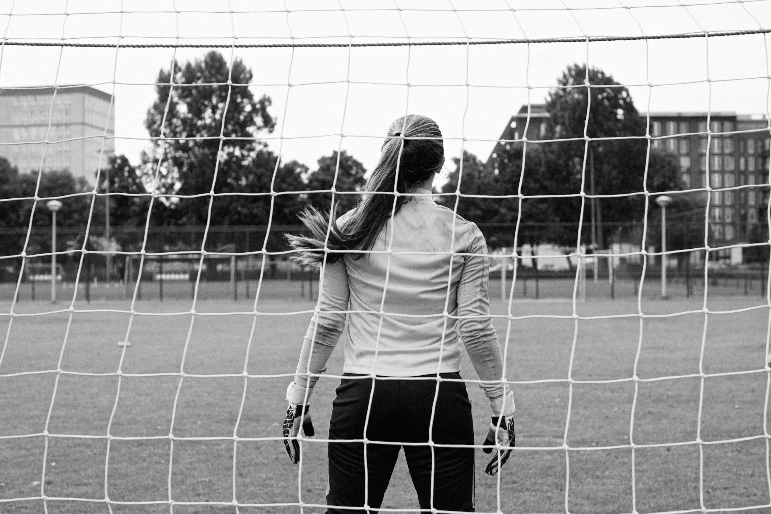 A female soccer goalie standing in front of the goal, looking onto the field, with trees and buildings in the background, in black and white.