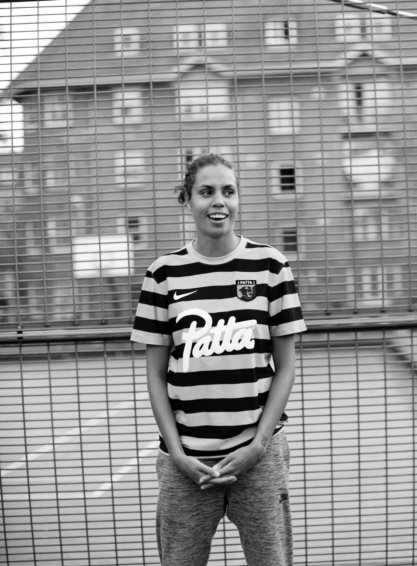 A woman standing in front of a wire fence, wearing a striped sports jersey and athletic pants, smiling and looking to the side.
