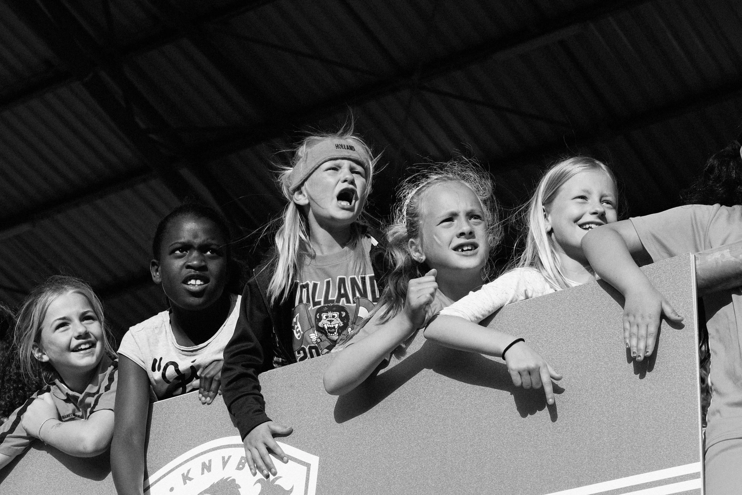 Group of young girls with excited and intense expressions, leaning on a barrier in what appears to be a stadium or sports arena.