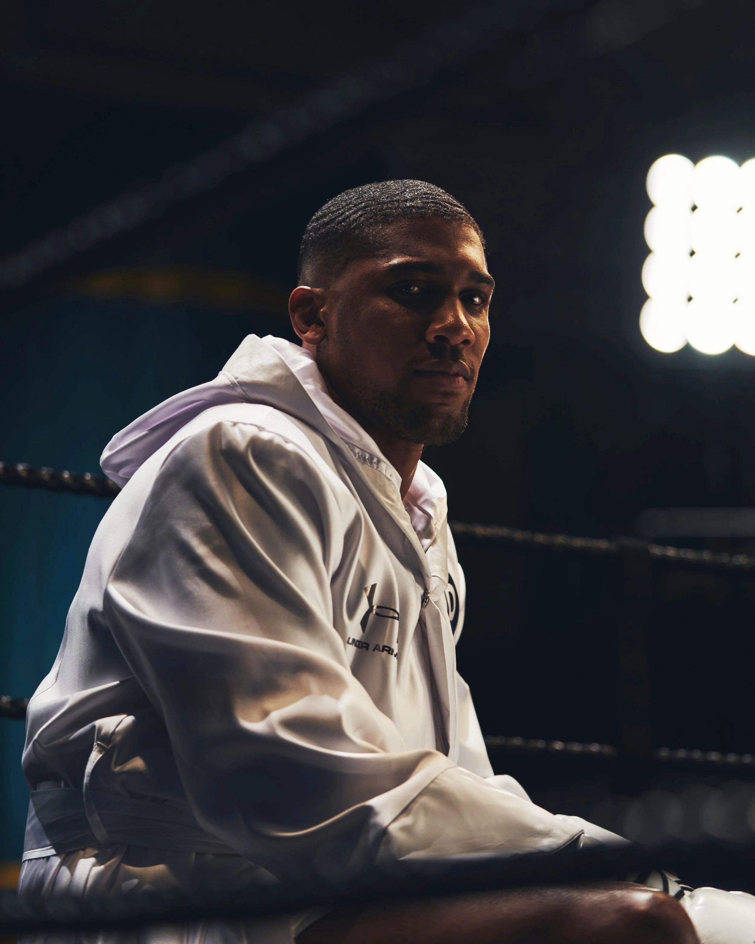 A man sitting inside a boxing ring, wearing a white jacket, with a serious expression on his face.