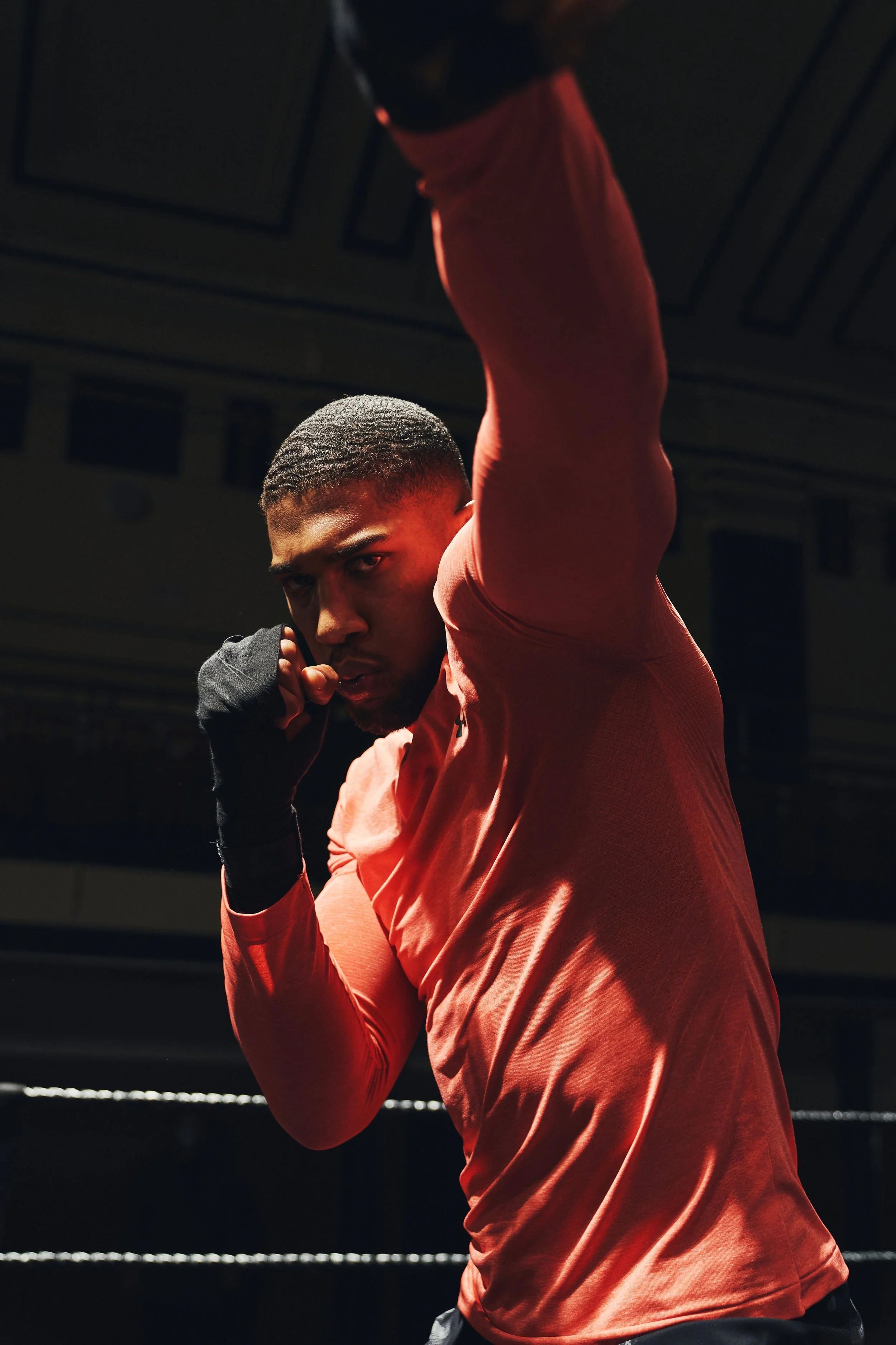 A male boxer in a red shirt and black gloves training in a boxing ring, with his left fist near his face and his right arm raised.