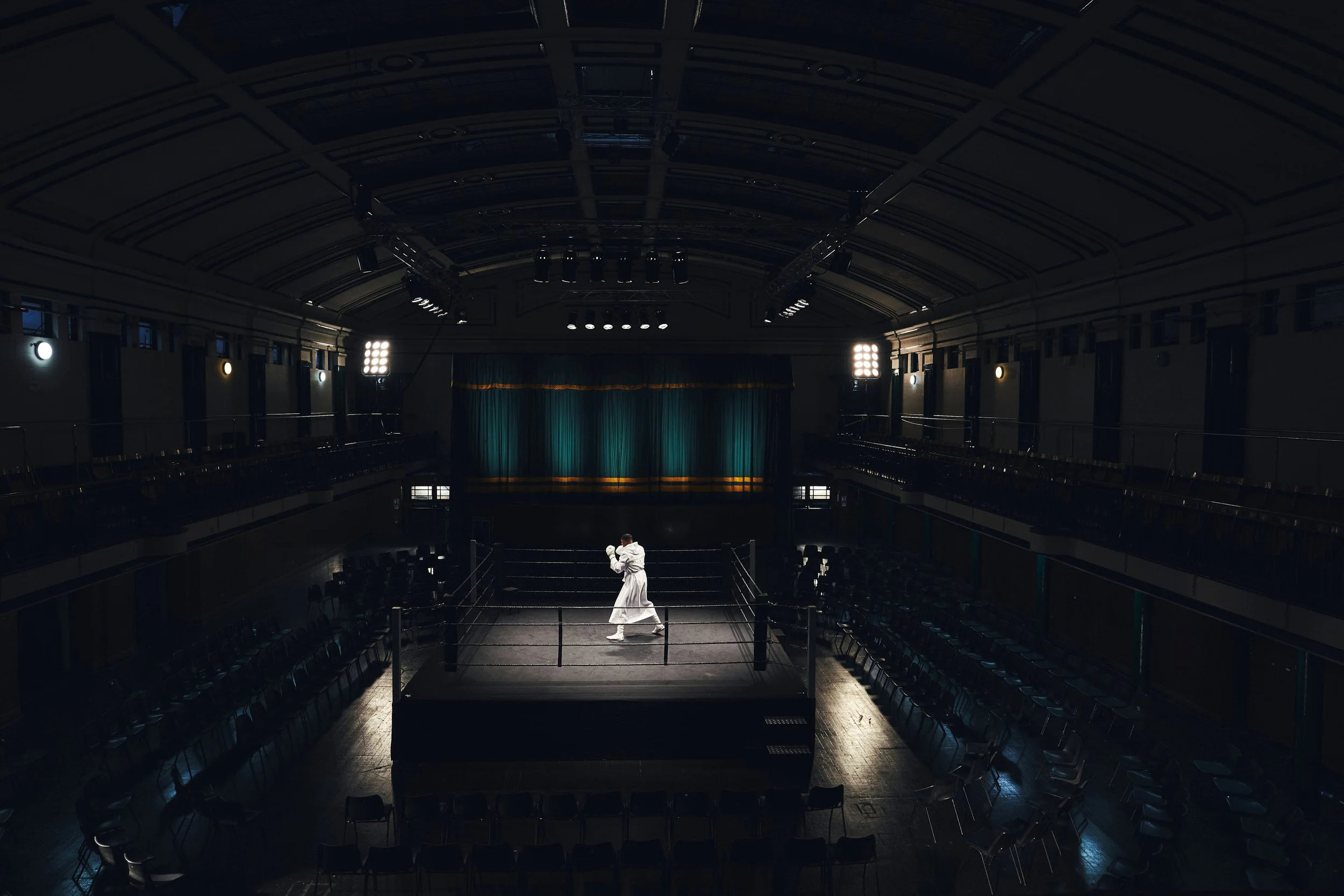 A person in a white jumpsuit practicing boxing inside an empty boxing ring in a dimly lit auditorium