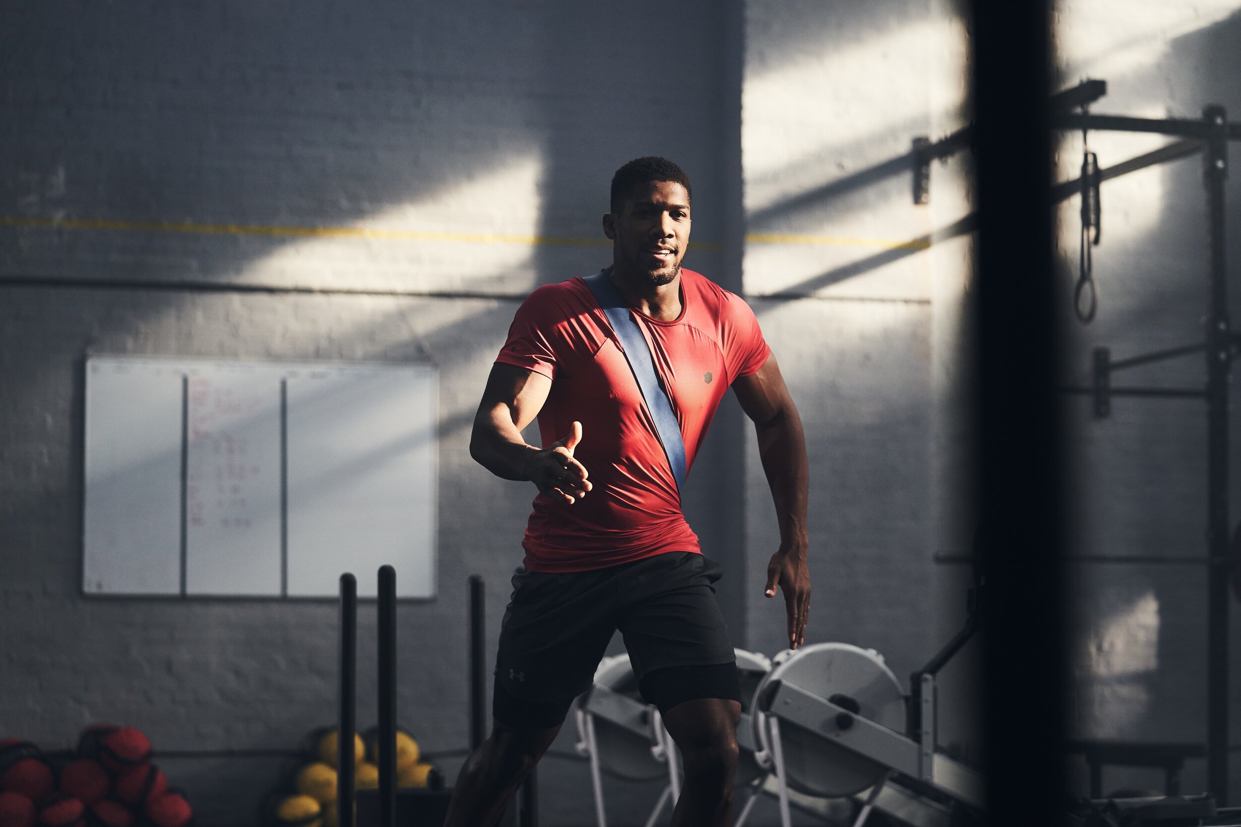A man in a red athletic shirt and black shorts running in a gym with workout equipment and a whiteboard in the background.