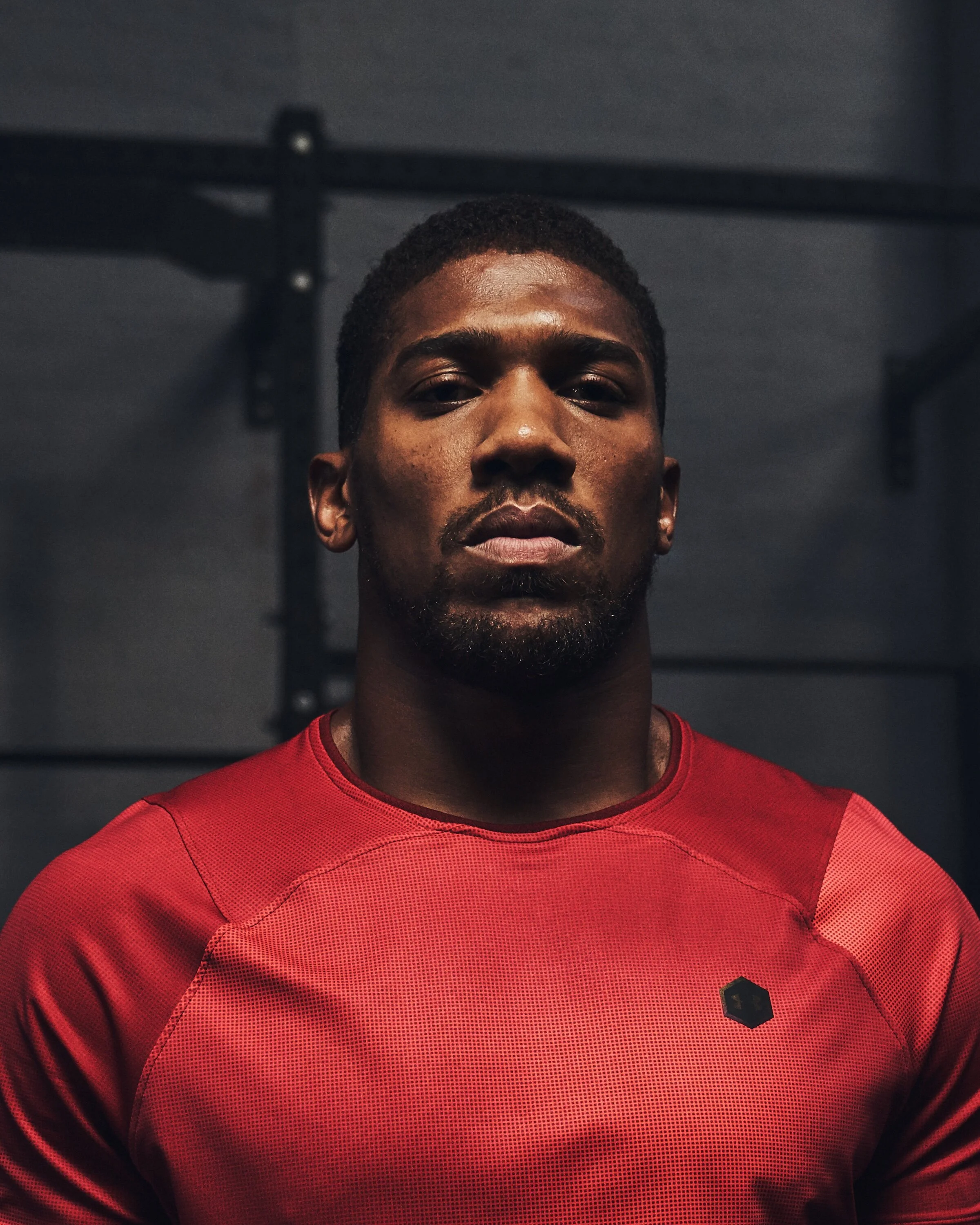 A man with dark skin wearing a red athletic shirt, standing indoors with a serious expression.