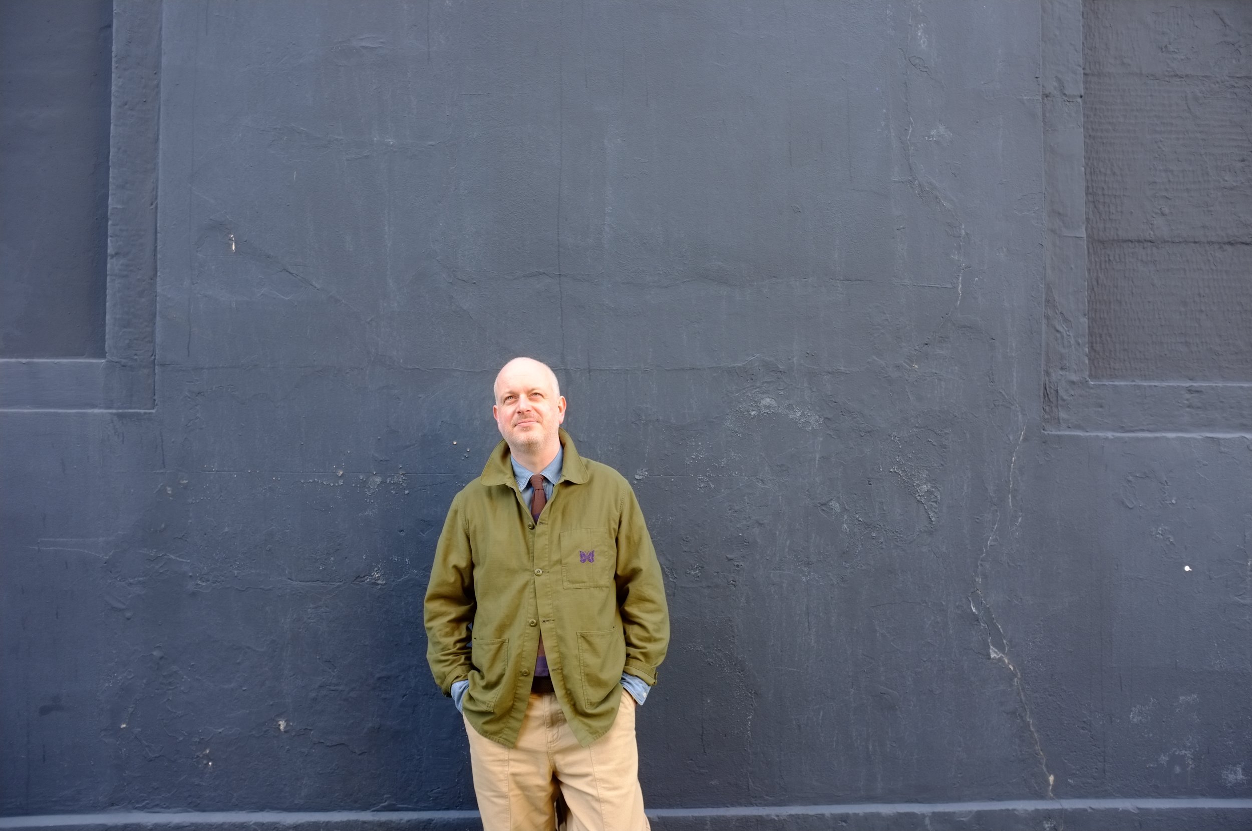Steven Doherty, Edinburgh-based brand marketing leader & consultant, wearing a green coverall jacket, denim shirt and striped tie, standing against a grey wall.