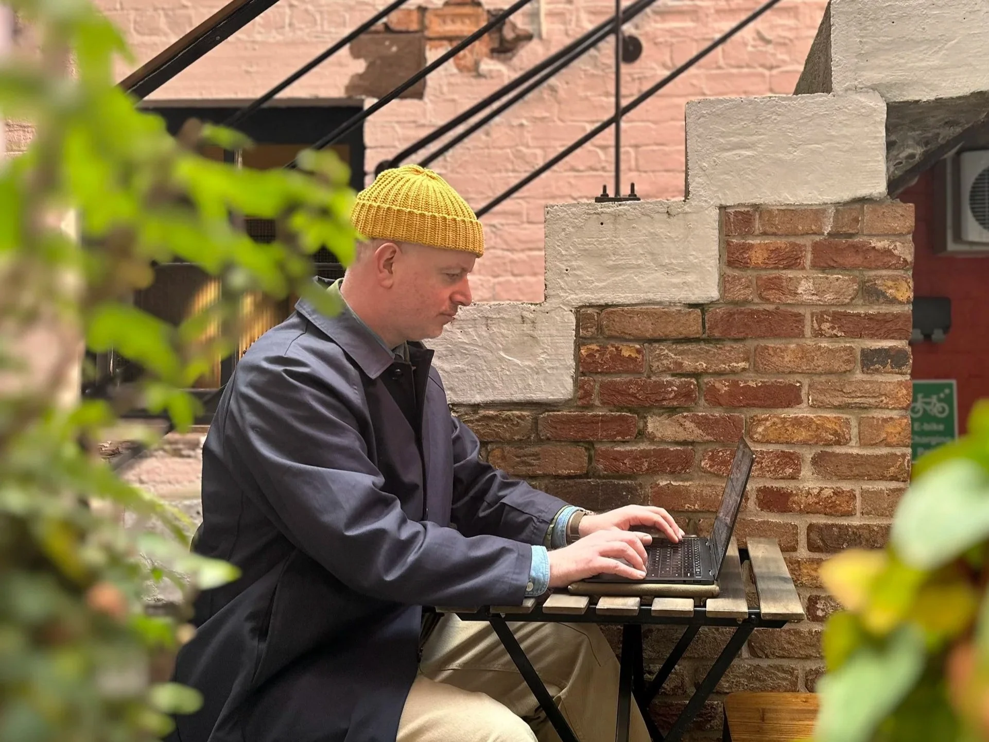 Steven Doherty, Edinburgh-based brand marketing leader & consultant, works outdoors on a laptop, wearing a navy coat, yellow knitted hat, beside stairs, near a painted brick wall, with some foliage in shot but out of focus.