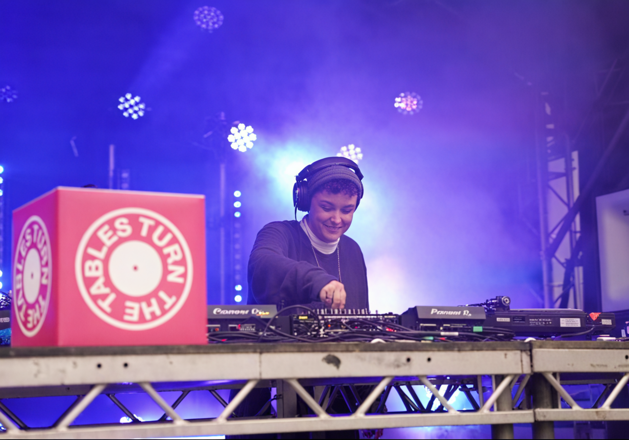 A female DJ wearing headphones and a black jacket, smiling while mixing music at a concert stage with colorful lights and fog, featuring a pink box with the text 'The Fabules Turn The'