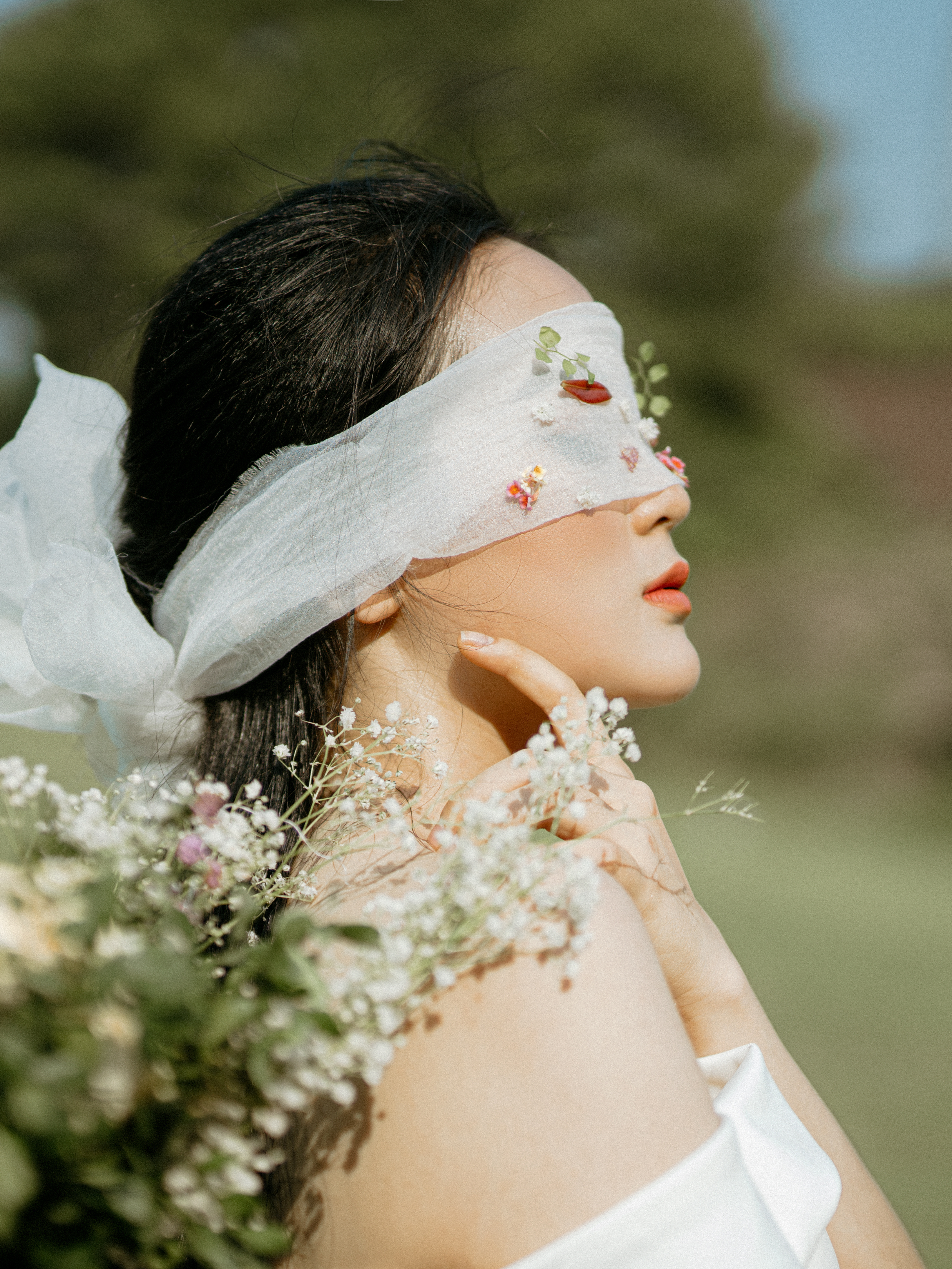 Une femme portant un bandeau blanc empêchant la lumière de son œil, entourée de fleurs blanches, avec un fond flou de verdure et de ciel bleu.
