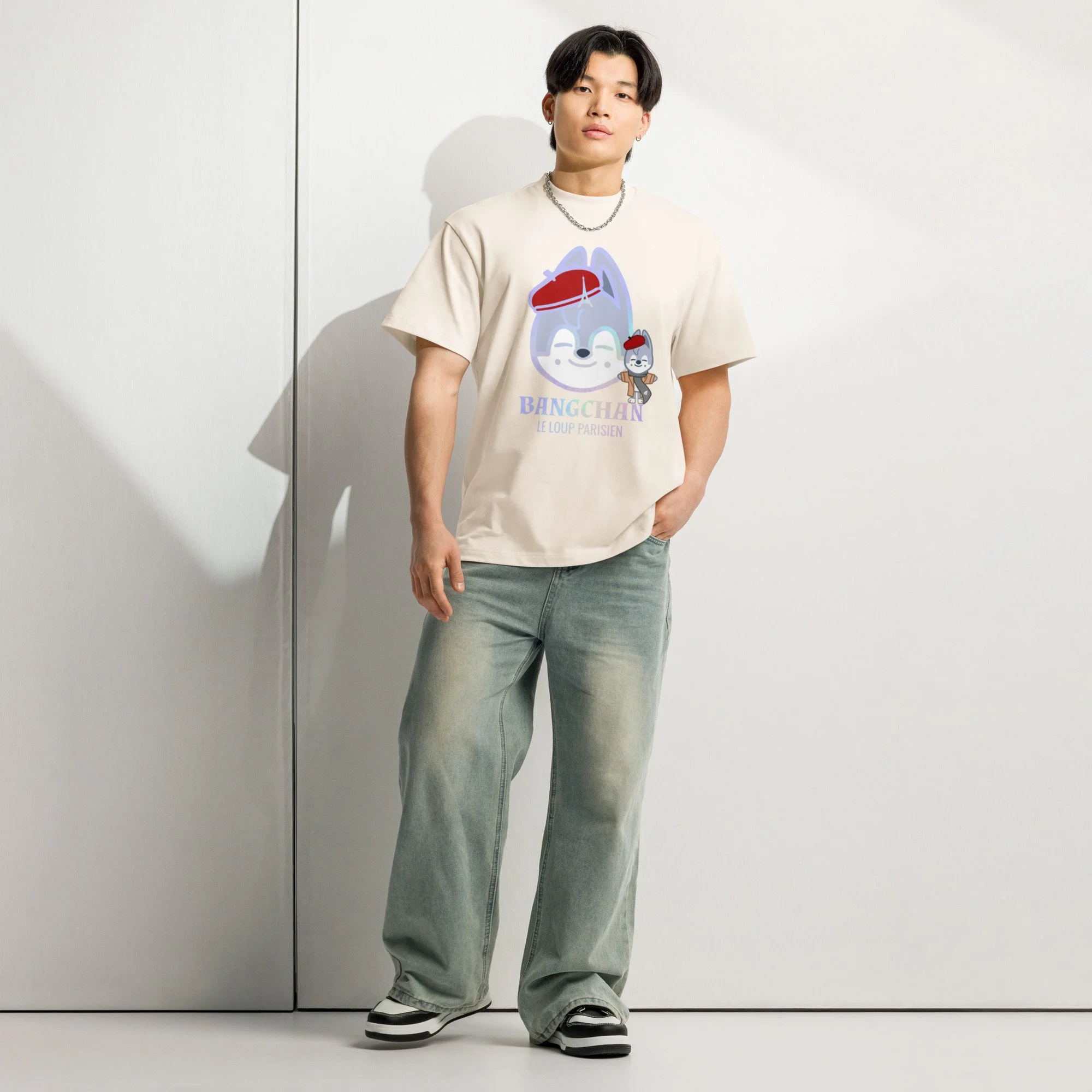 Jeune homme debout contre un mur blanc, portant un t-shirt avec un dessin de tête de loup stylisée, des jeans larges, des chaussures de sport et une chaîne en argent.
