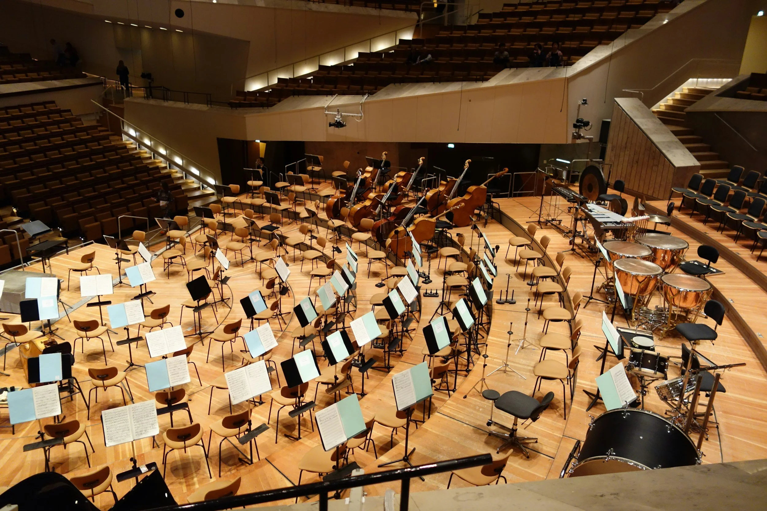 Une salle de concert avec des sièges en bois, des instruments de musique (piano, violons, timbales, etc.) disposés sur scène, et un éclairage tamisé.