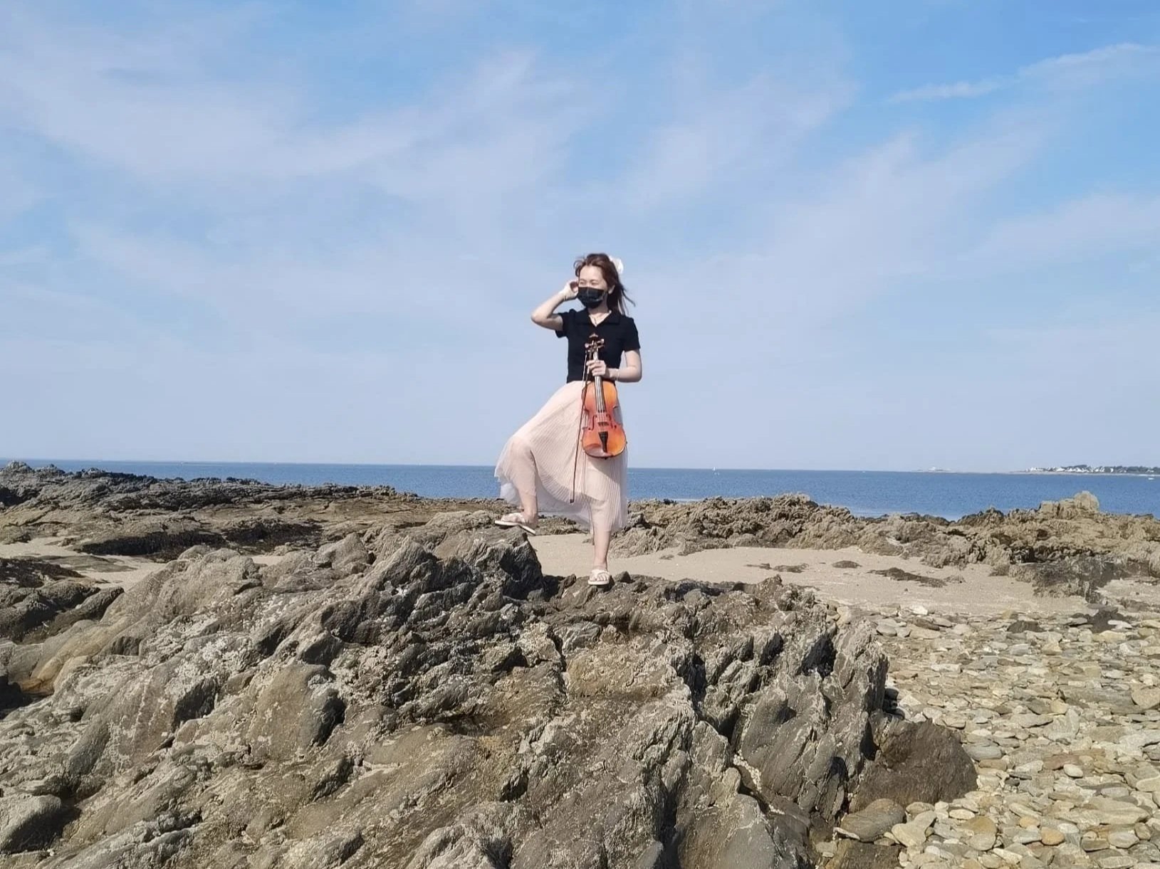 Une femme jouant du violon sur une plage rocheuse, portant un masque noir, une robe longue beige, et un haut noir, face à la mer avec un ciel bleu clair.