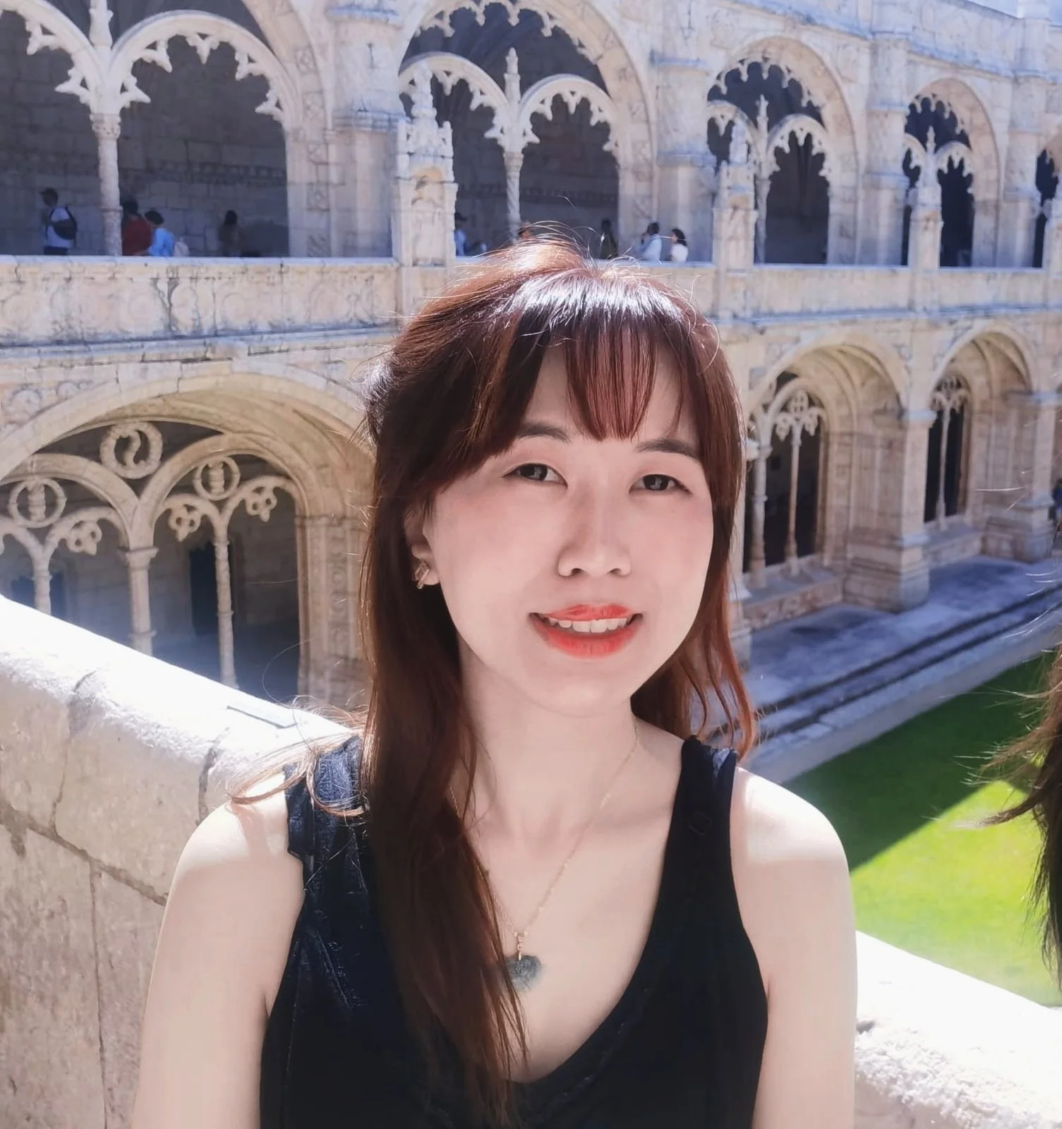 Jeune femme avec longs cheveux bruns, souriante, portant une robe noire, posant devant un bâtiment historique en pierre avec des arches et des colonnes, en plein jour.