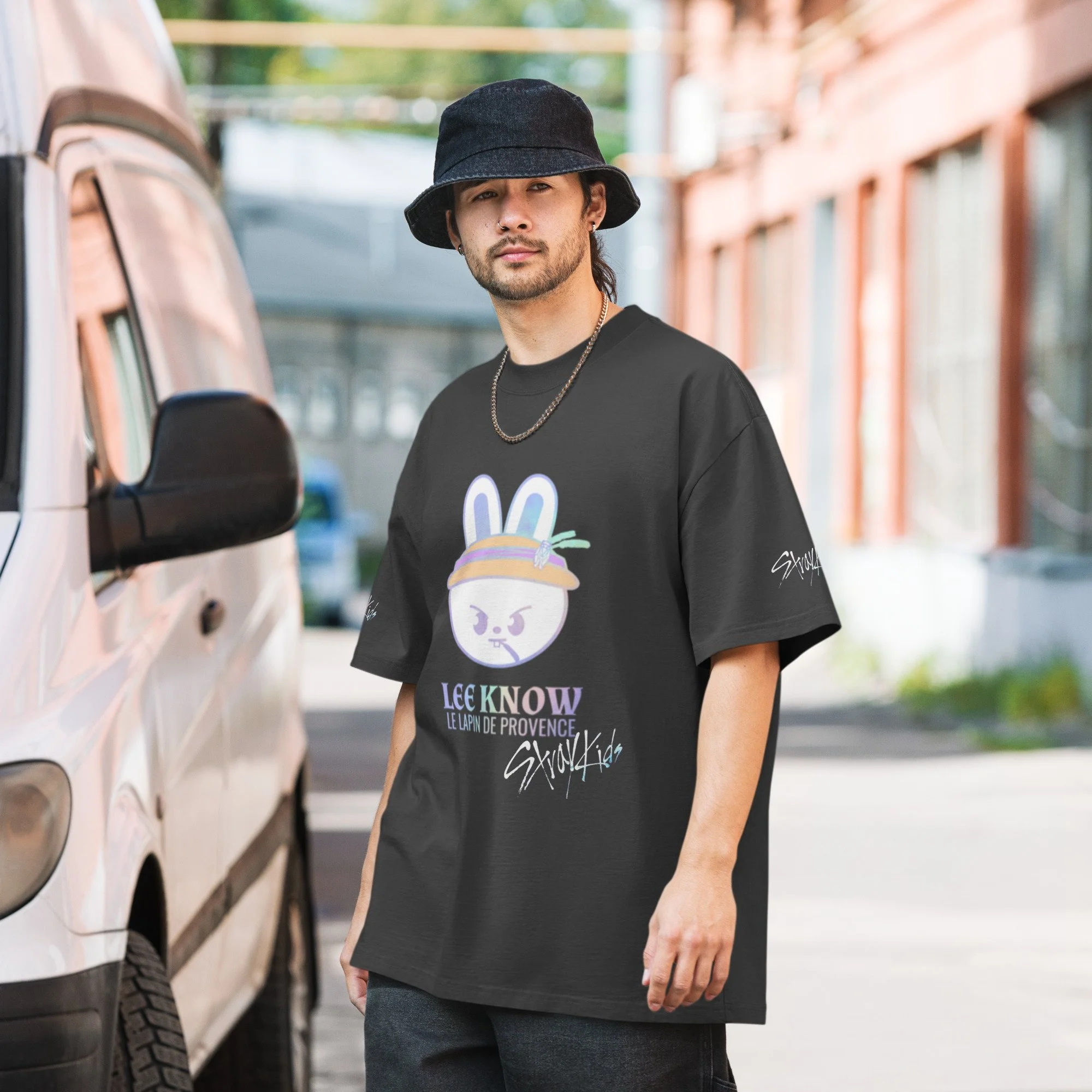 Un jeune homme portant un chapeau noir, un t-shirt noir avec un dessin de lapin, une chaîne en argent, se tient à côté d'un véhicule blanc dans une zone urbaine.