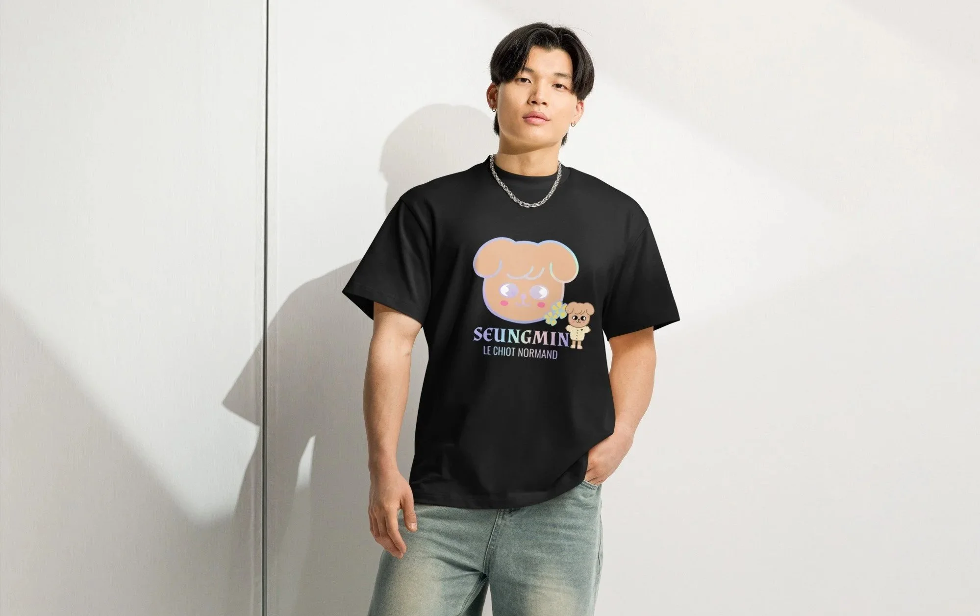 Un homme portant un t-shirt noir avec un dessin d'un chien et le texte "Seungmin Le Chiot Normand" se tient contre un mur blanc, avec une chaîne en argent autour du cou, regardant vers l'appareil photo.