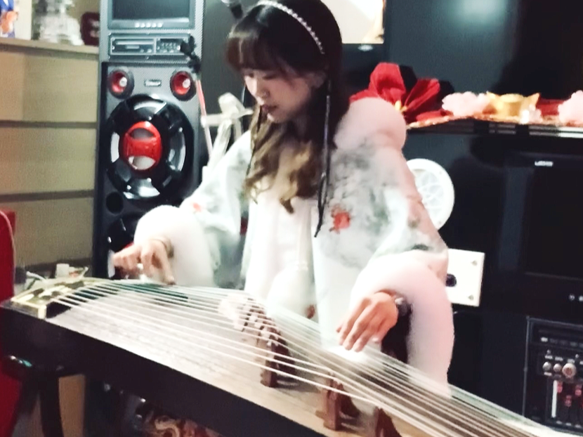 Une femme joue du guzheng, un instrument de musique chinois, dans une pièce comme un salon ou un studio, avec un haut-parleur et un appareil audio en arrière-plan.