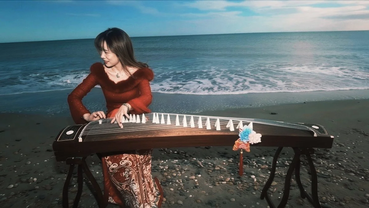 Une femme joue d'une guzheng sur une plage en bord de mer, vêtue d'une robe rouge