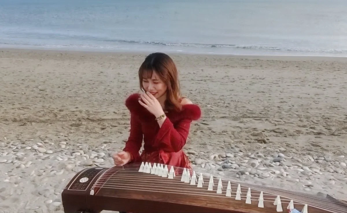 Une femme souriante jouant du guqin sur une plage sableuse, avec la mer en arrière-plan.