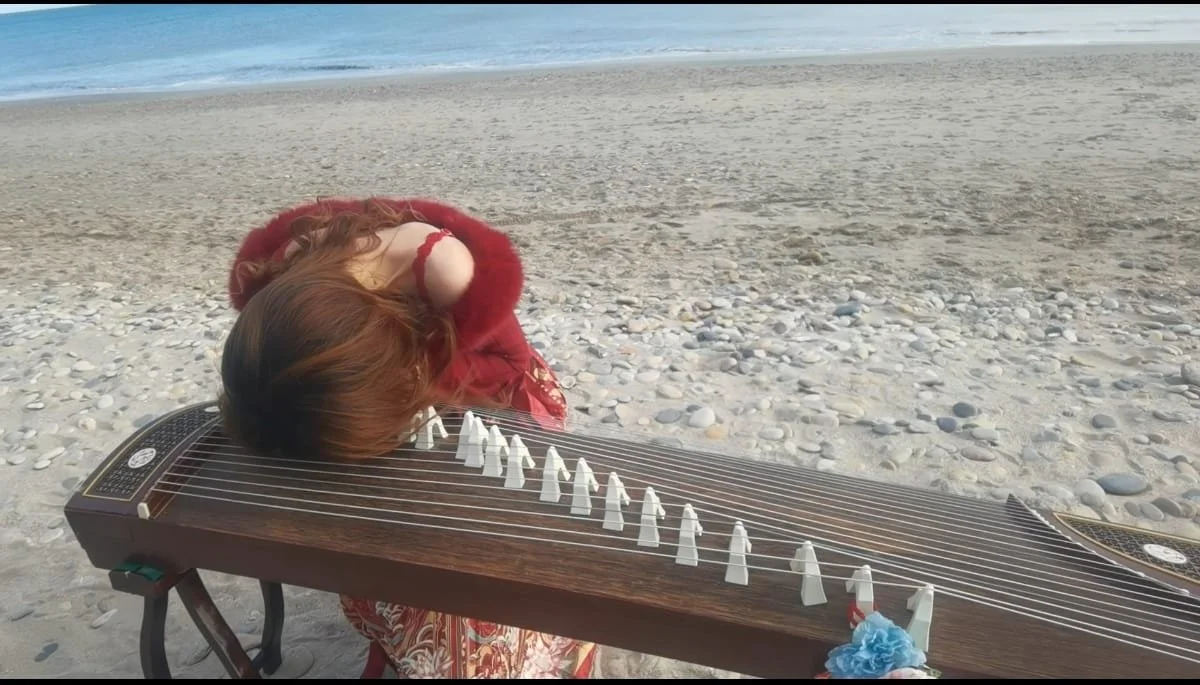 Une femme jouant de la cithare sur une plage de sable. La mer est visible en arrière-plan.