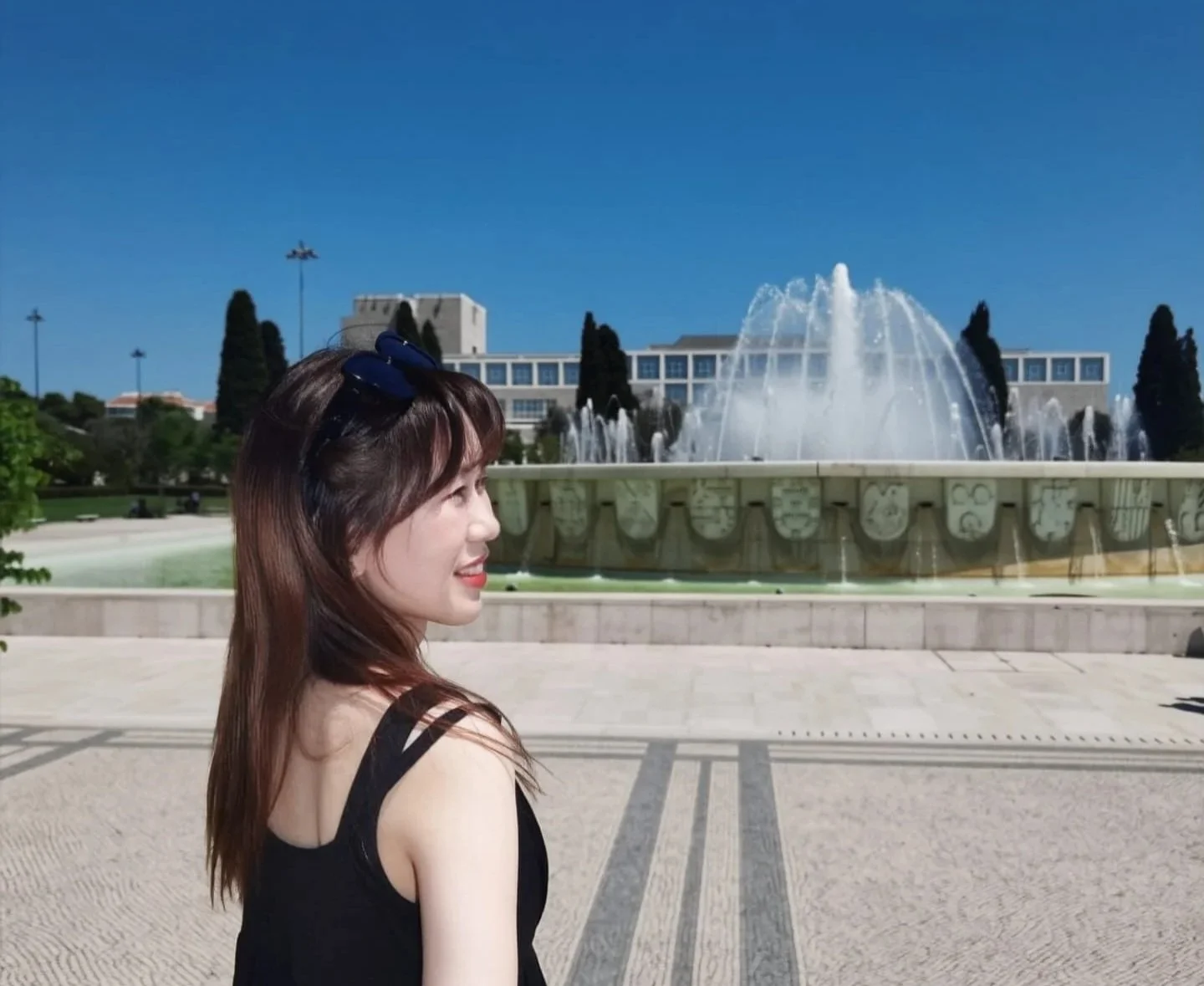 Femme souriant devant une fontaine en plein air avec bâtiment moderne en arrière-plan, ciel bleu clair.