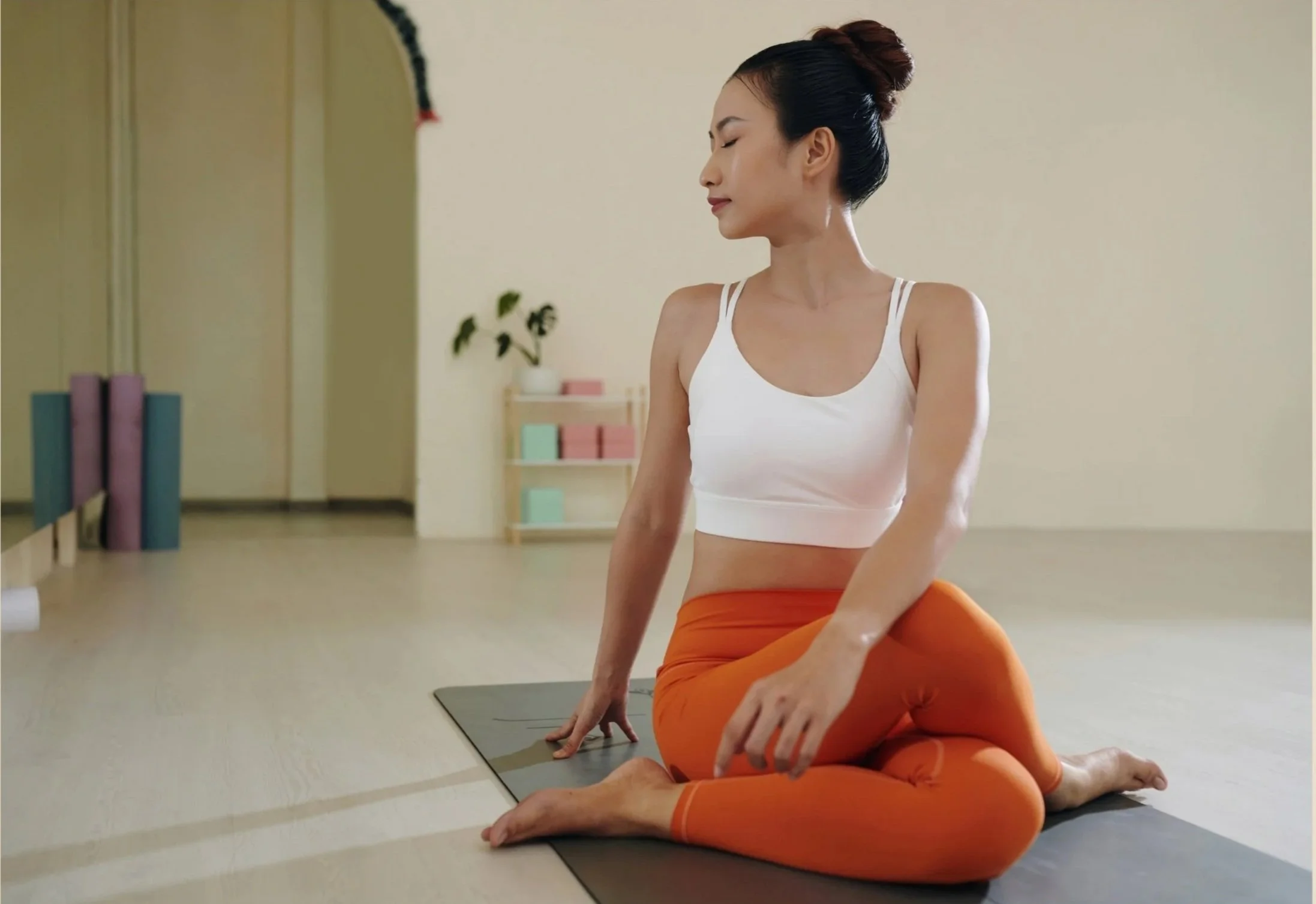 Une femme pratique le yoga en position assise sur un tapis de sol dans une salle claire, avec des étirements et une expression de concentration.