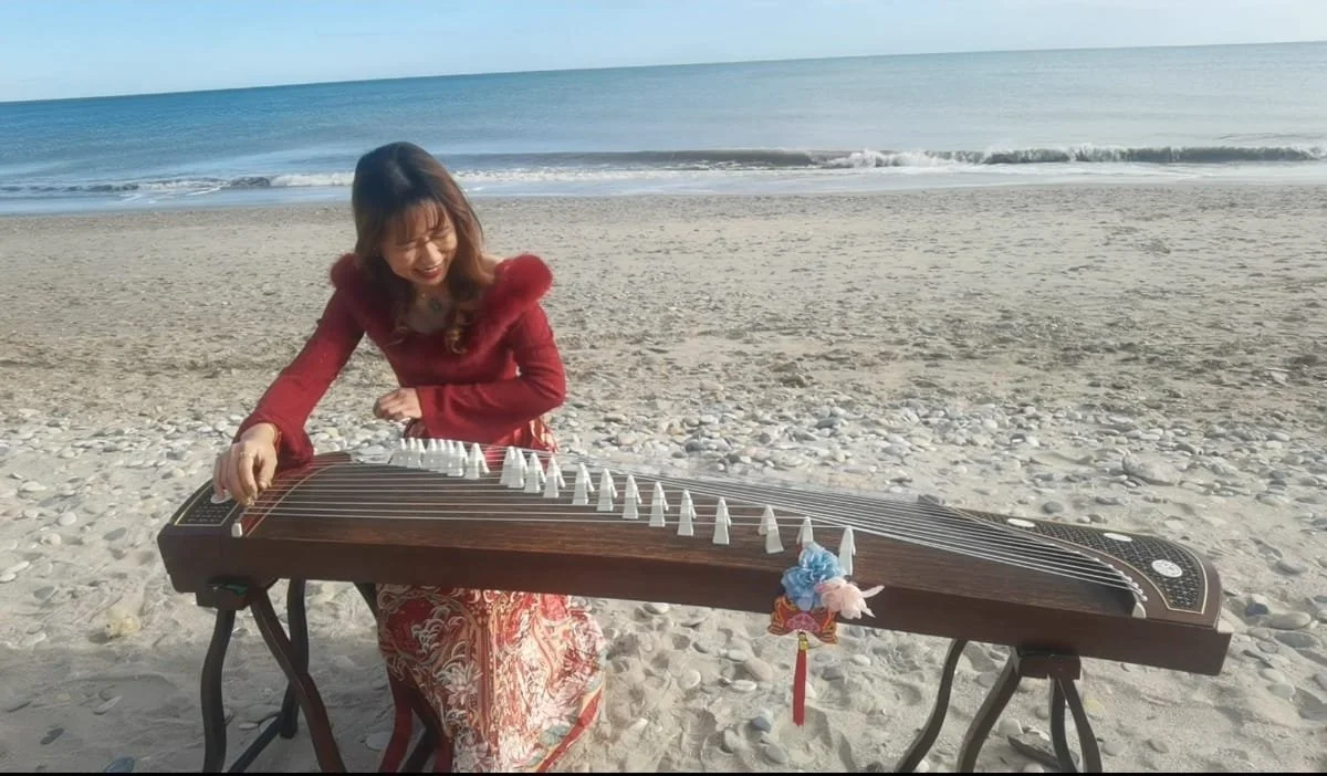 Une femme joue du guzheng sur la plage, avec la mer en arrière-plan, portant une robe rouge et un manteau rouge avec une capuche en fourrure