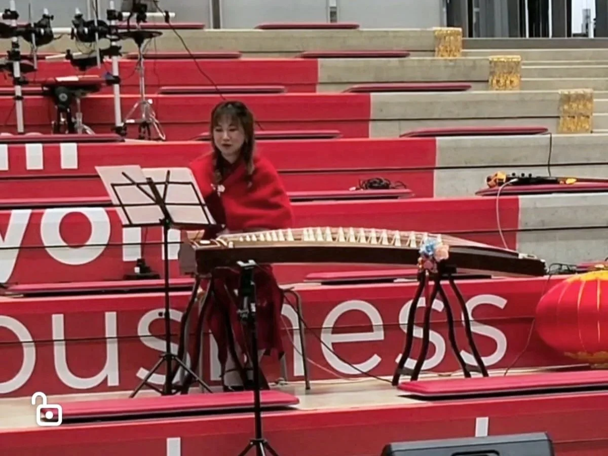 Femme jouant du guzheng, un instrument de musique chinois, dans un espace décoré de rouge avec des gradins. 