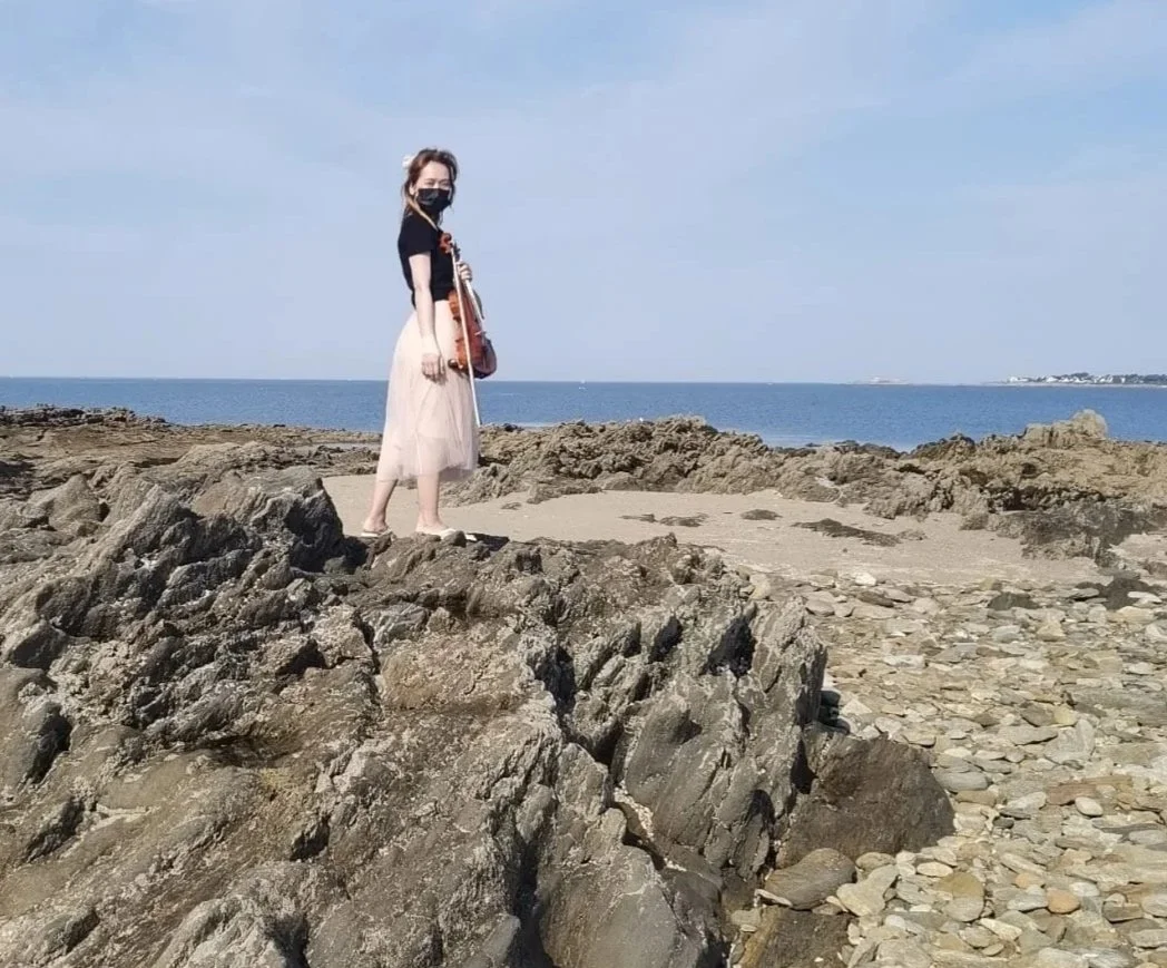 Jeune femme portant un masque noir, une robe beige et un haut noir, debout sur des rochers au bord de la mer, face à l'océan, par une journée ensoleillée.