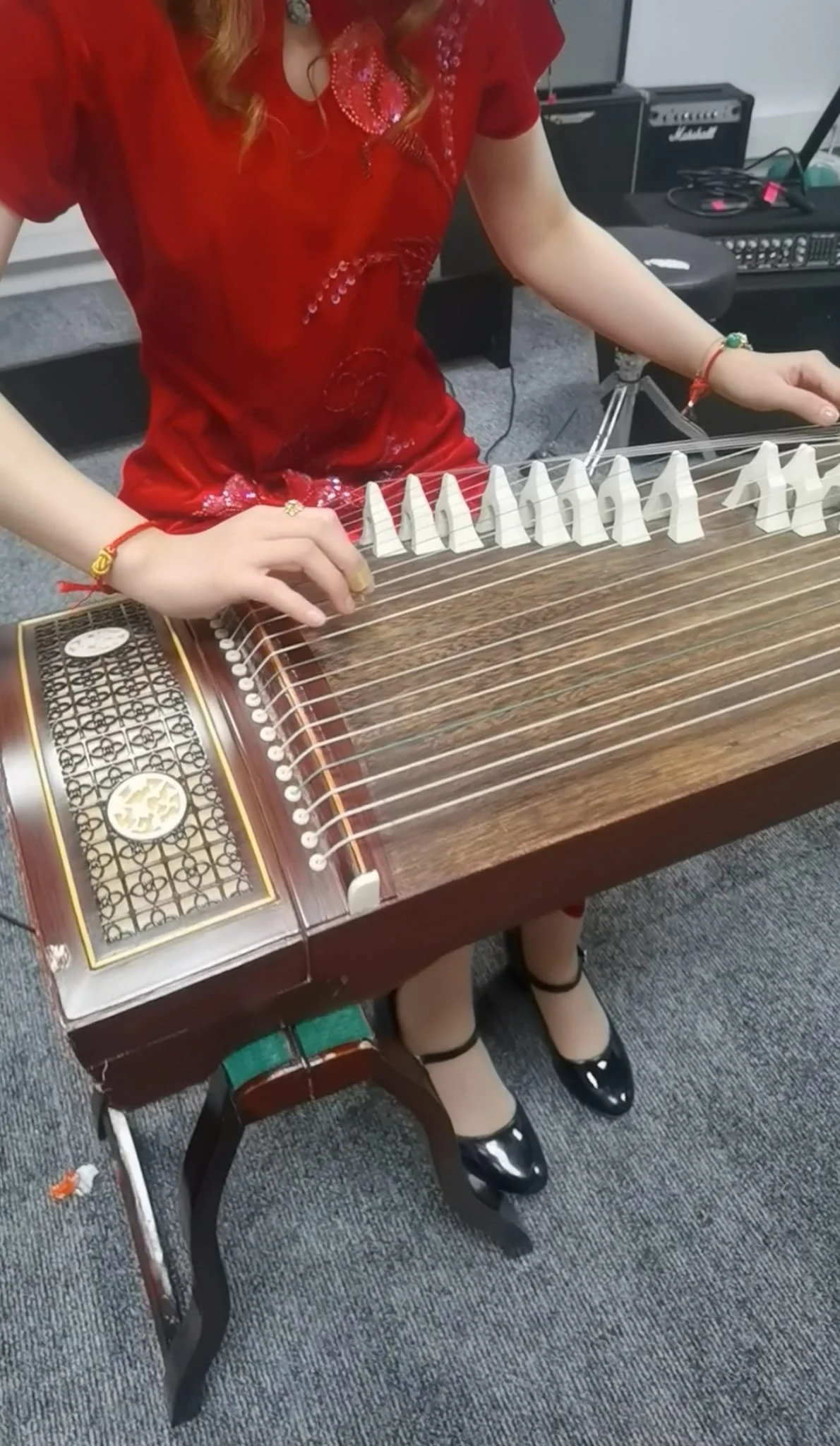Femme assise jouant du guzeng, instrument de musique traditionnel chinois, portant une robe rouge avec des bijoux, chaussures noires vernies, sur un sol en tapis gris.