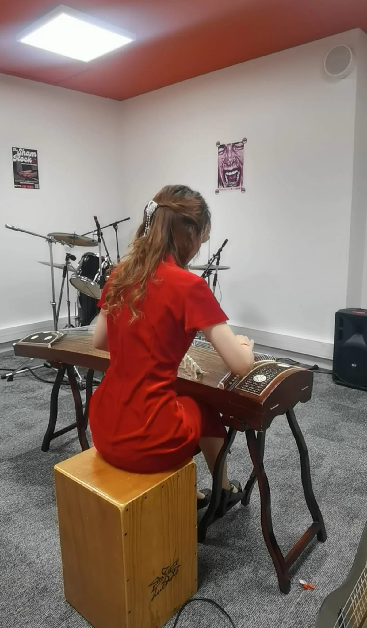Une femme s'exerce à jouer du koto dans une pièce, avec une batterie en arrière-plan et un microphone sur la droite, portant une robe rouge, assise sur un tabouret en bois, face à une table de jeu.
