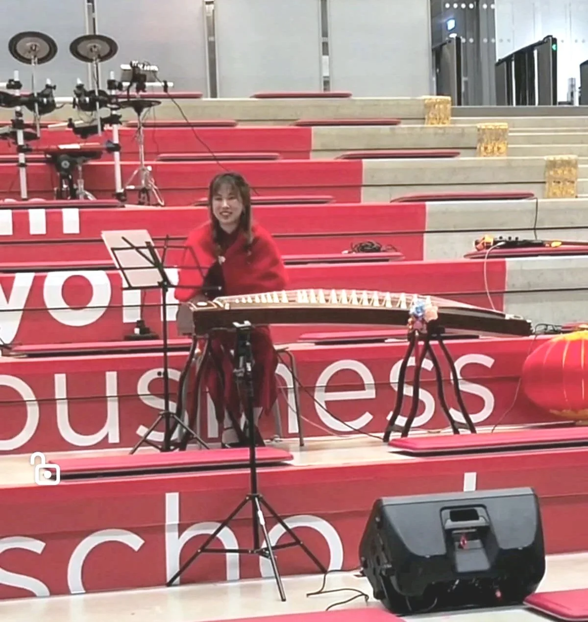 Une jeune femme jouant du guzhENG sur un podium en intérieur, avec des escaliers en arrière-plan, décoré de rouge et blanc.